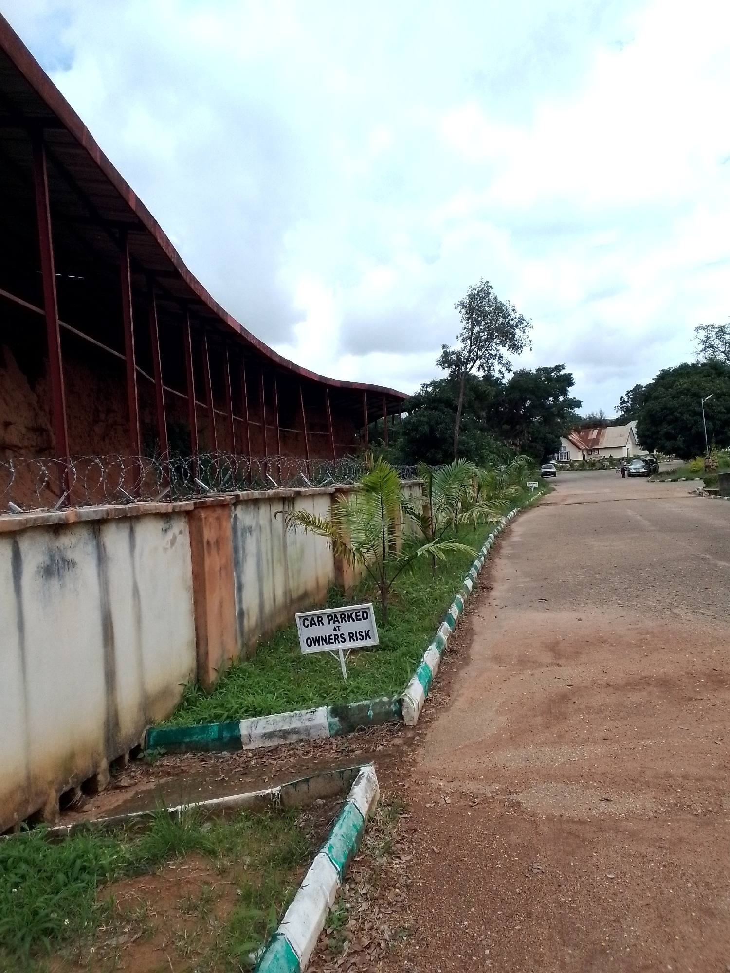 The image depicts an outdoor scene with a road curving to the right, leading towards a building complex. The road surface appears to be a mix of asphalt and dirt. On the left side of the road, there is a weathered concrete wall topped with barbed wire fencing, behind which a large, reddish, curved structure with a metal roof is visible. Small palm-like plants are growing along the wall. A white sign with black lettering reading "CAR PARKED AT OWNERS RISK" is planted in the grass near the wall. In the distance, several parked cars are visible on the road, and two figures are walking. The background consists of lush green trees and a cloudy sky. The presence of the barbed wire and the "owners risk" sign suggests a potentially unsecured or hazardous parking area. The architectural style of the buildings and the vegetation point towards a tropical or sub-tropical climate. Based on the information available, this scene is located in the city of Jos, Nigeria.