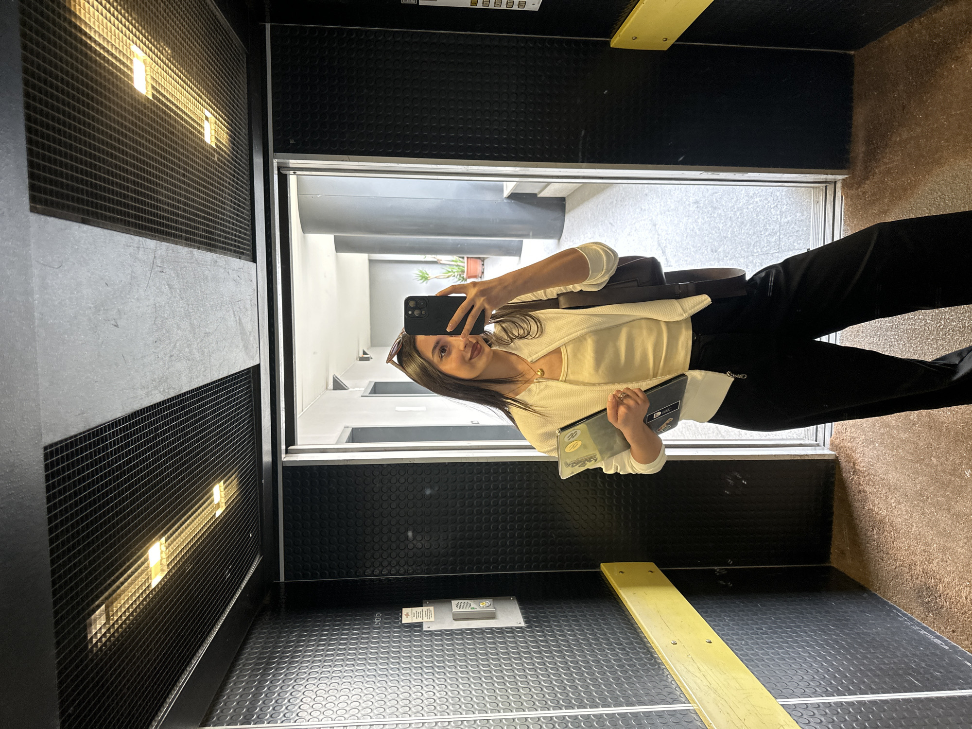 A young woman takes a selfie in an elevator. She holds a smartphone in her right hand and a book/passport in her left. The woman wears sunglasses on her head, a white cardigan, and black pants. The elevator has black textured walls and a yellow handrail. The door is open, revealing a hallway with a potted plant. The elevator is located in Milan, Italy.