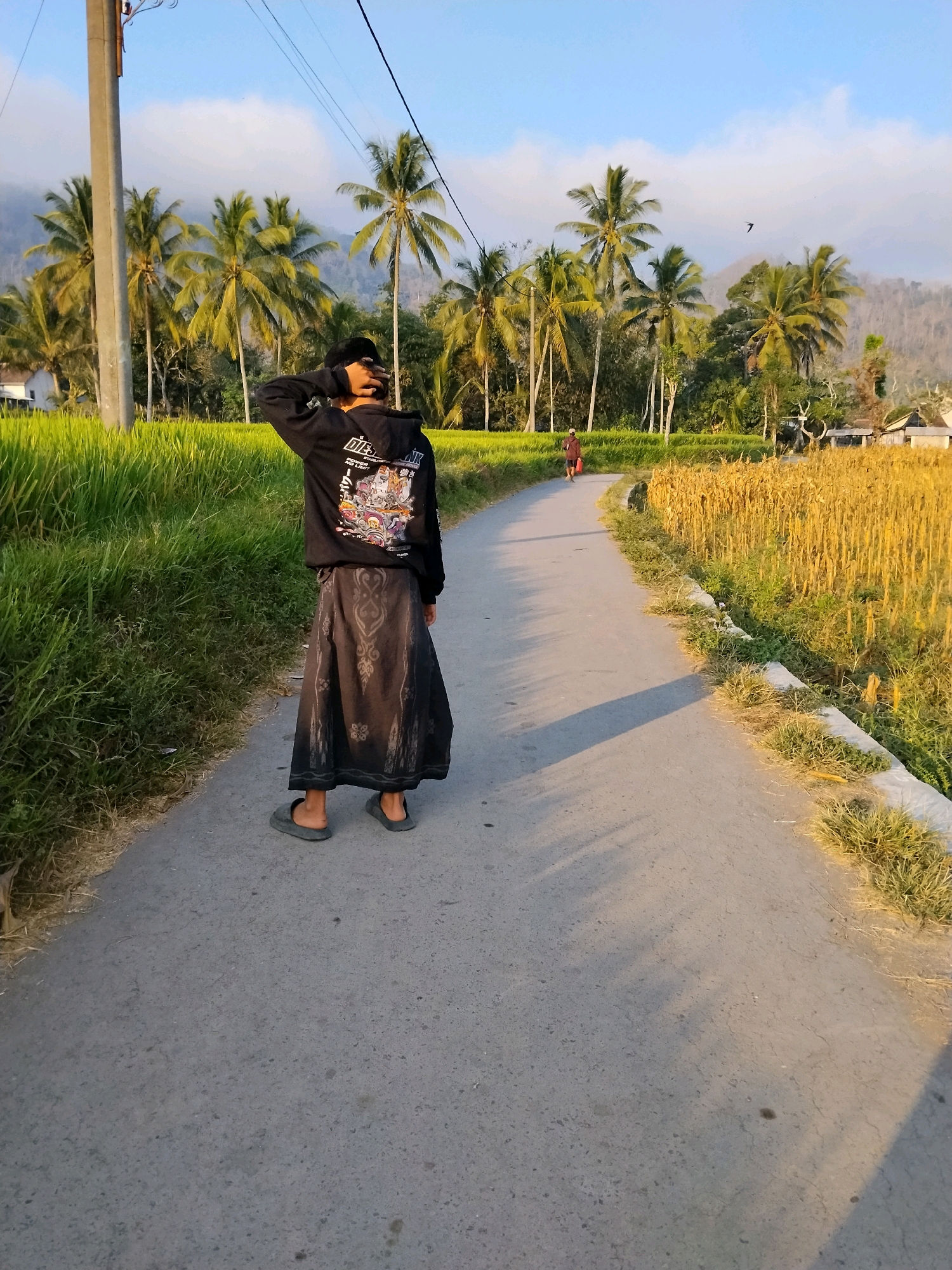 A young person wearing a black hoodie, a dark patterned skirt, and grey flip-flops stands facing away from the camera on a paved road. Their left arm is raised, and they are touching the back of their head. The road curves to the right and stretches into the distance. On the left side of the road, there is a vibrant green field of rice paddies bordered by several palm trees. On the right side of the road, there is a field of dry, golden stalks, likely corn, with a low concrete border lined with grass. In the background, more palm trees and dense vegetation are visible, along with some houses and a hazy, mountainous landscape under a pale blue sky with scattered clouds. Another person is walking in the distance on the road, carrying a red bag. A bird is flying in the sky. The lighting suggests it is late afternoon, with long shadows cast across the road. The scene depicts a rural setting in or near Tlogosari, Indonesia.