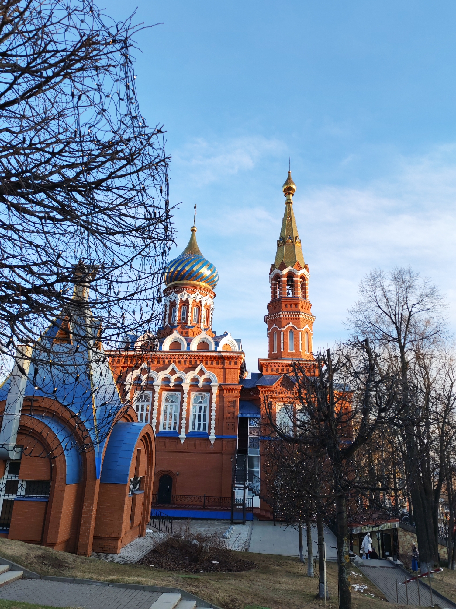 The image depicts the Holy Michael Cathedral in Izhevsk, Russia, under a bright blue sky with scattered clouds. The cathedral's distinct architecture features red brick construction with white and blue accents. Two prominent towers rise above the main structure; one has a blue and gold striped dome, and the other is a tall, slender spire topped with a golden spire. Bare tree branches, some adorned with small lights, frame the cathedral. A smaller, blue-roofed building is connected to the main cathedral structure. The landscape slopes downward toward the right side of the frame, where a paved walkway leads to what appears to be an entrance to the cathedral grounds. Several individuals are walking on this path. The scene conveys a clear, daytime view of this religious landmark within its urban setting.