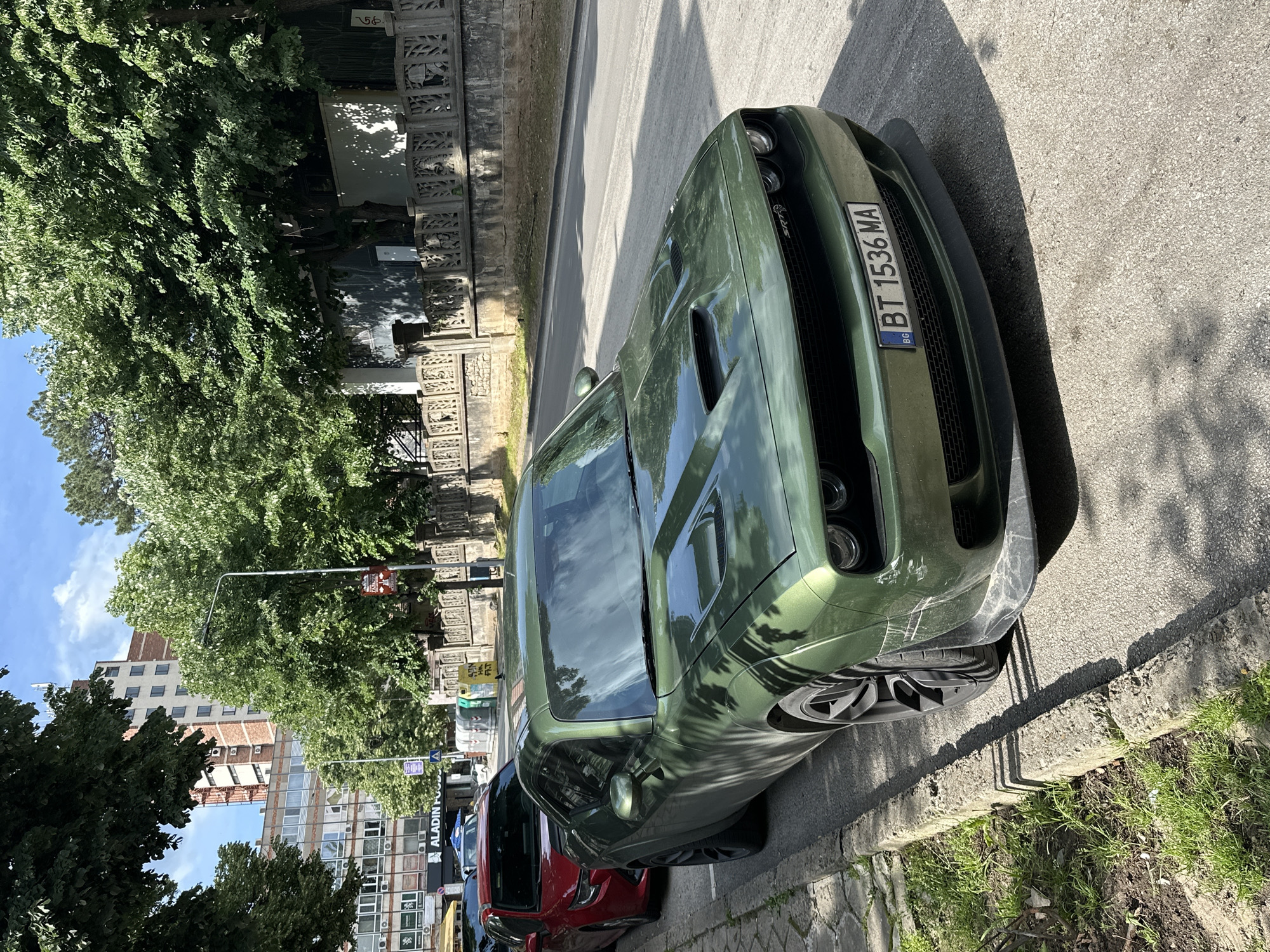 The image shows a green Dodge Challenger SRT parked on a street in Veliko Tarnovo, Bulgaria. The car has a Bulgarian license plate, "BT 1536 MA." It appears to be parked legally on the side of the road, next to a curb with grass and weeds growing along it. There are other cars parked behind the Dodge, one appears to be red. Buildings and trees are visible in the background. The image is taken from a slightly elevated position, looking down at the car. The front of the car shows damage. The building in the background has a sign "Aladin Food".
