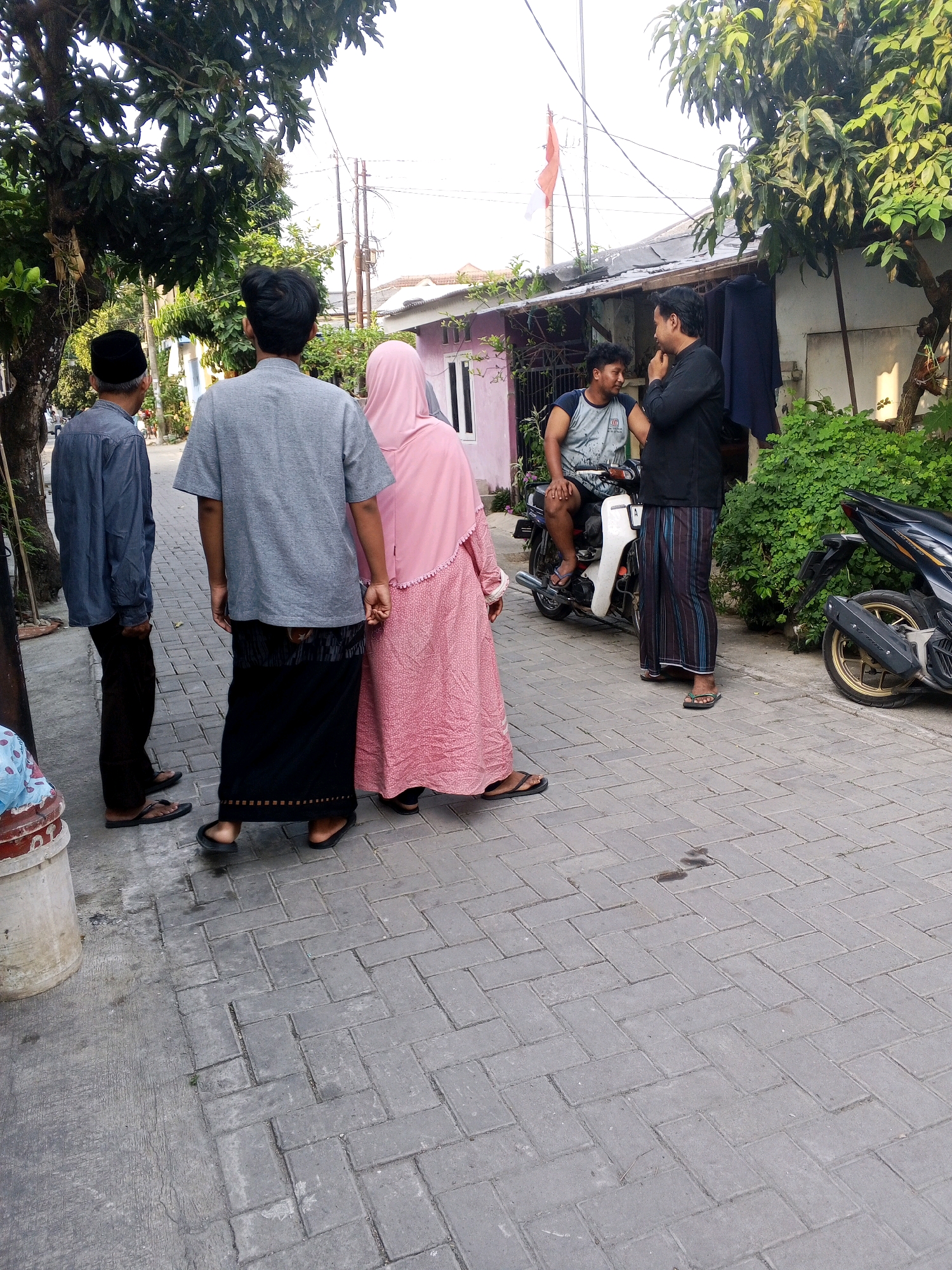 The image depicts a street scene in Ciruas, Indonesia, under a bright sky. In the foreground, three people are visible from the back. A man on the left wears a blue long-sleeved shirt, dark pants, and black flip-flops. Beside him, a man in a grey t-shirt and a black sarong with a patterned border, also wearing black flip-flops. To his right, a woman in a pink hijab and a long pink dress, wearing black flip-flops, holds his hand.
In the middle ground, two men are engaged in conversation. One is seated on a white motorcycle, wearing a light blue and grey t-shirt and dark shorts. The other man stands next to him, wearing a black long-sleeved shirt and a striped sarong, with his hand on his chin. A dark blue garment hangs from a rack behind them. To the far right, a black motorcycle is parked, its gold-rimmed wheel visible.
The environment includes lush green trees providing shade, a building with a weathered roof and pink walls, and power lines overhead. The ground is paved with grey interlocking bricks. A large, light-colored cylindrical object, possibly a bin, is partially visible in the lower left corner. A red and white Indonesian flag is mounted on a pole in the background. The overall atmosphere suggests a casual outdoor gathering or interaction.