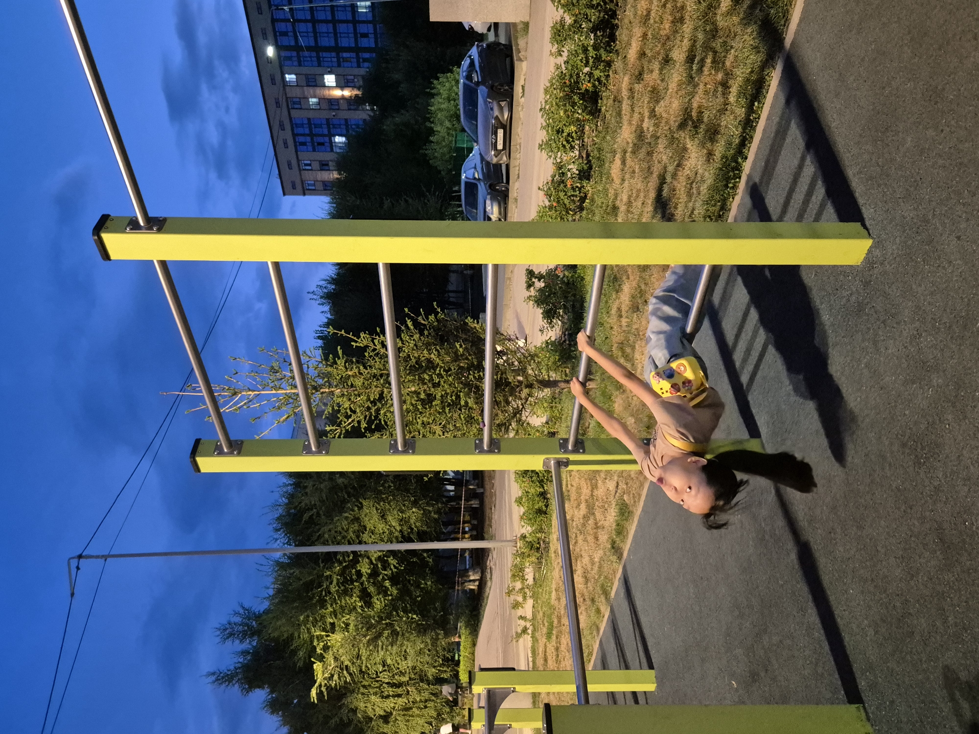 The image captures a child on a horizontal bar at a street workout area in Kokshetau, Kazakhstan. The child, a young girl with long dark hair, is hanging upside down, with her arms gripping the bar. She is wearing a beige t-shirt and light blue jeans. The workout equipment is painted bright yellow with silver metal bars. The ground beneath the equipment is a dark grey rubberized surface, bordered by dry grass. In the background, there are trees, a street with cars parked, and a multi-story building with illuminated windows. The sky is a deep blue, indicating it is dusk.
