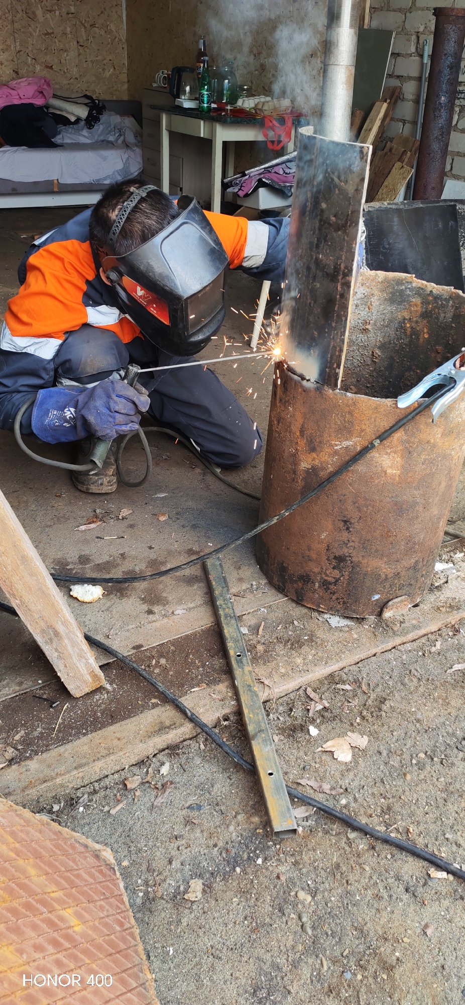 A person wearing protective welding gear, including a mask and gloves, is welding a metal object outdoors in Chaikovsky, Russia. The person is kneeling on a concrete surface and using a welding torch to join a vertical metal bar to a cylindrical metal object. Welding sparks are visible. Several other metal objects are present, including a long bar and a flat piece of metal. A table, some bottles, and a bed can be seen in the background.