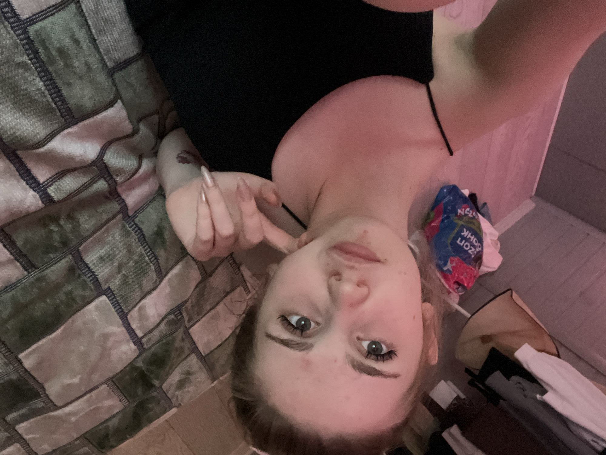 The image shows a young woman lying on a bed. She is wearing a black top and her arm is raised, with her fingers near her face. A tattoo is visible on her arm. The bed is covered with a patterned blanket in shades of green and pink. In the background, there is a blue plastic bag with text that appears to be in Cyrillic, and some folded clothing. The lighting suggests it may be evening or indoors with artificial light, casting a pinkish hue on the wall. The woman's expression is neutral, and she is looking towards the camera. The scene appears to be a casual, personal setting, likely a bedroom. The location context provided is Ashkadar, Russia.