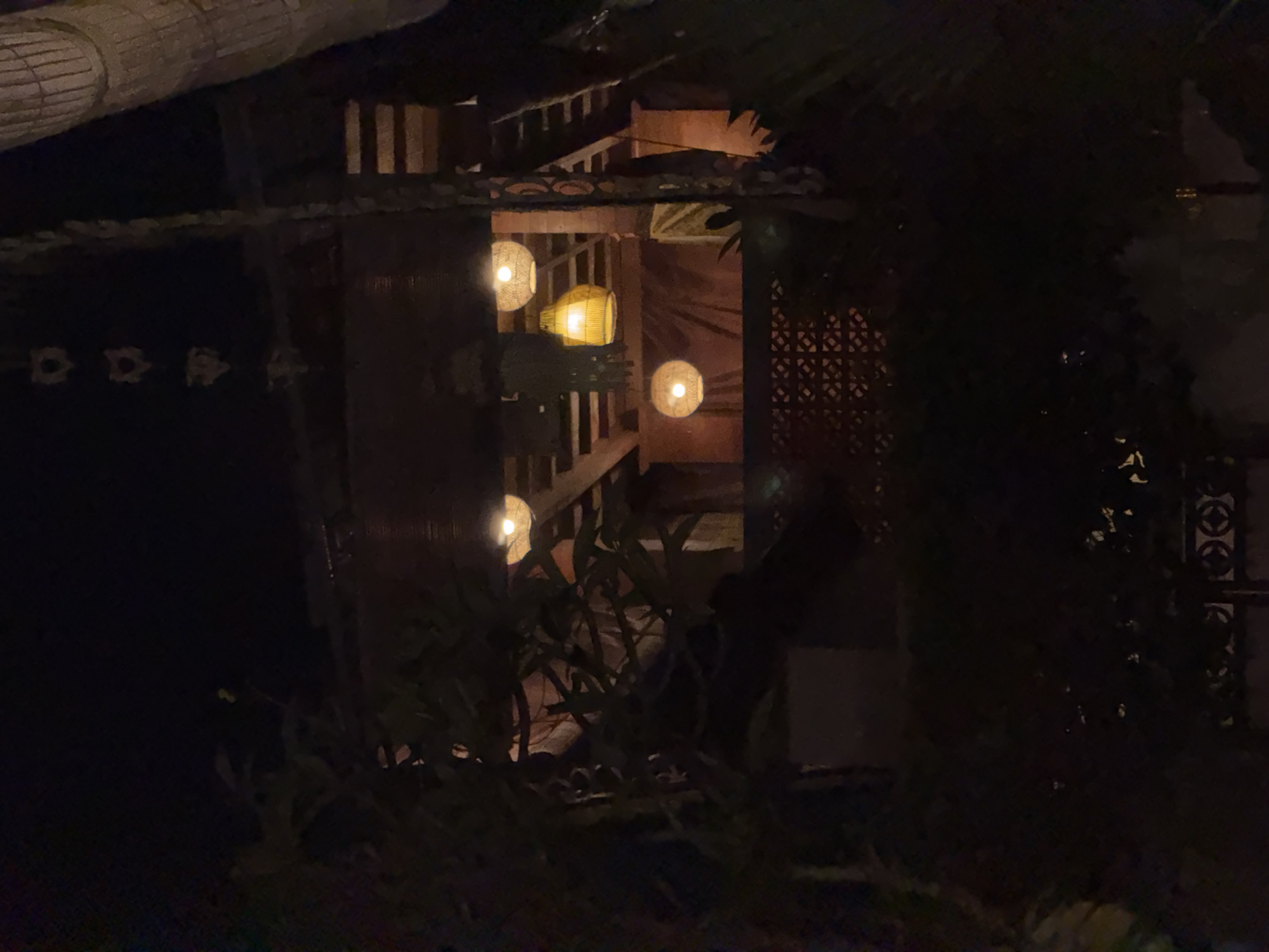This media file depicts a dimly lit scene at night, likely in Sukawati, Indonesia. The central focus is a wooden structure with multiple levels, possibly a balcony or an open-air dining area, adorned with several spherical lanterns. These lanterns emit a warm, yellow glow, illuminating the immediate surroundings and creating a cozy, inviting ambiance.
The structure features wooden railings and vertical supports, suggesting traditional Indonesian architecture. Some of the lanterns are made of woven material, hinting at a rustic or artisanal design. Lush green foliage is visible in the foreground, partially obscuring the view and adding a natural element to the scene. The overall impression is one of tranquility and peacefulness, characteristic of a tropical evening.
The darkness of the background indicates it is night, and the absence of any specific weather elements suggests a clear or calm night. The scene evokes a sense of relaxation and possibly dining or socializing in an outdoor setting. There is no visible text in the media file.