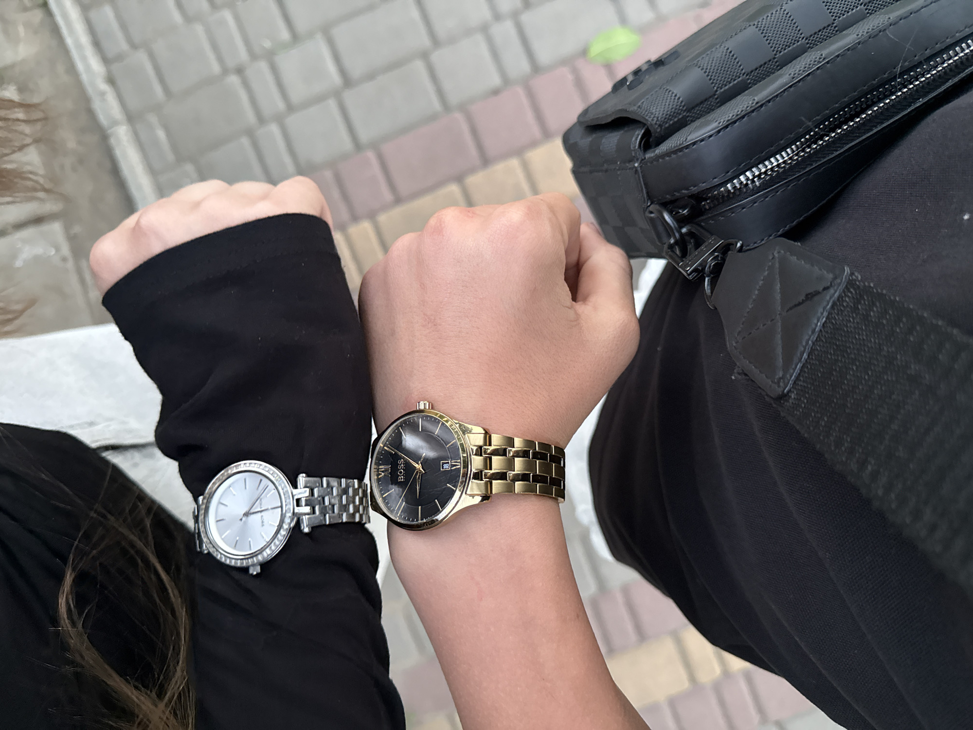 The image shows two wrists wearing watches. The wrist in the foreground wears a gold-toned wristwatch with a black dial, featuring roman numerals and a date window. The brand "BOSS" is visible on the dial. The wrist in the background wears a silver-toned wristwatch with a white dial and a diamond-encrusted bezel. Both individuals are wearing black clothing. A black checkered patterned bag is visible in the lower right corner. The background appears to be a paved walkway with square pavers. The image is taken outdoors.