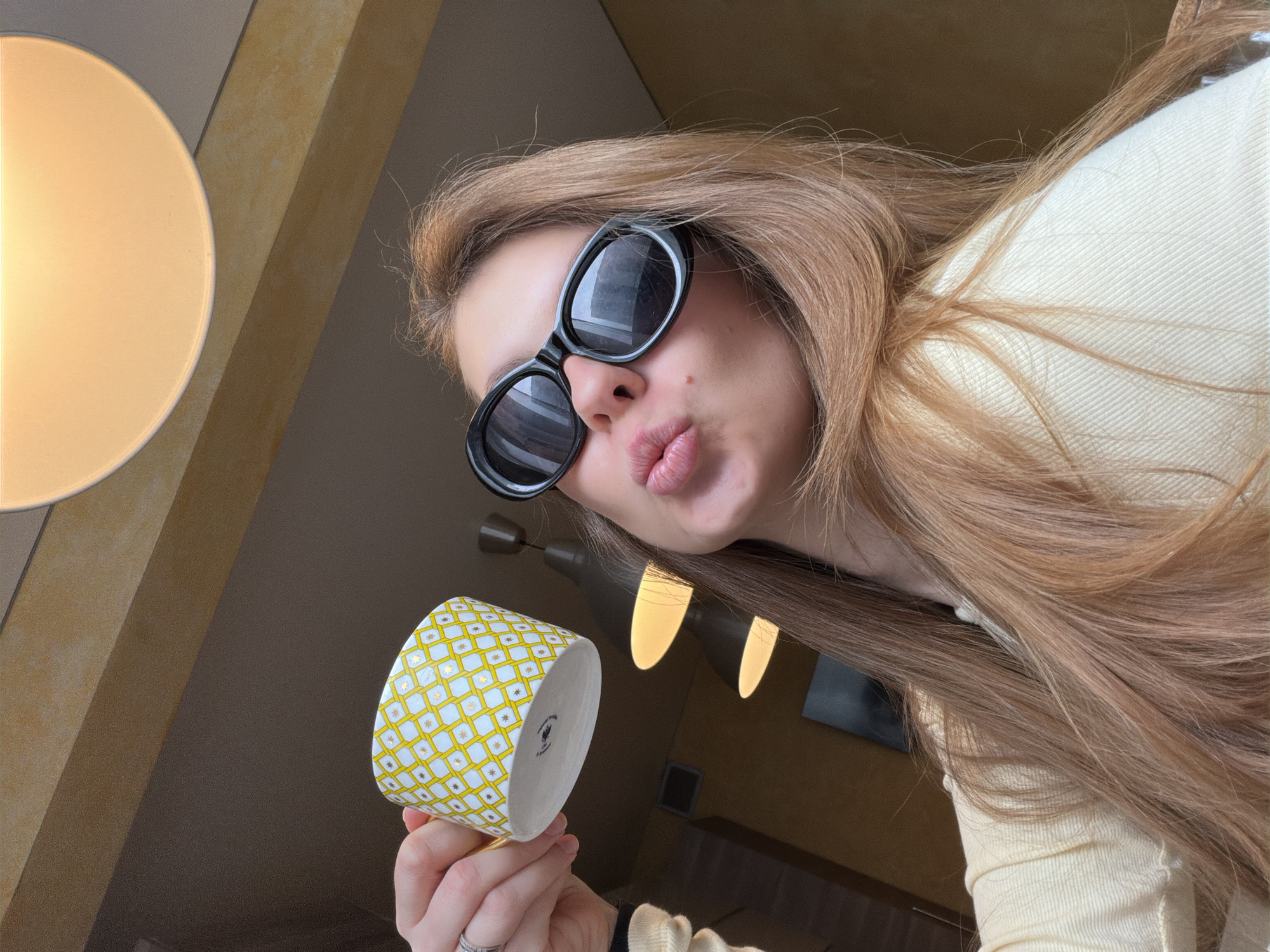 A woman with long blonde hair and wearing black sunglasses is holding a yellow and white patterned mug. She is making a kissing face at the camera. A round lamp is visible in the upper left corner of the image. The photo was taken in Khimki, Russia.