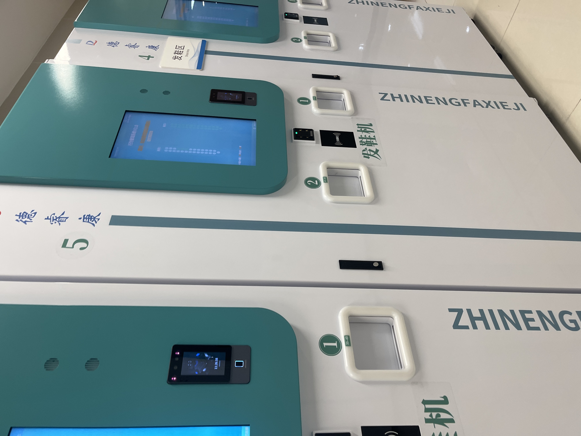 The image shows a row of white and teal automated dispensing machines, likely in a public facility. The machines are labeled with Chinese characters and numbers, suggesting they dispense items and are organized into different sections. One machine is labeled with the number "5" and Chinese characters that translate to "De Kang." Another machine is labeled "4" with "De Kang" and "Shoe Area." Below the screens on some machines are numbered compartments, such as "1" (with size "39-40") and "2" (with size "41-42"), for dispensing items. One machine has a clear indication of "发鞋机" which translates to "shoe dispensing machine." There is a facial recognition scanner on one of the machines. The setting appears to be indoors, with a tiled floor visible. The overall context suggests these machines are part of a service provided in a specific location within Yuzhong District, China.