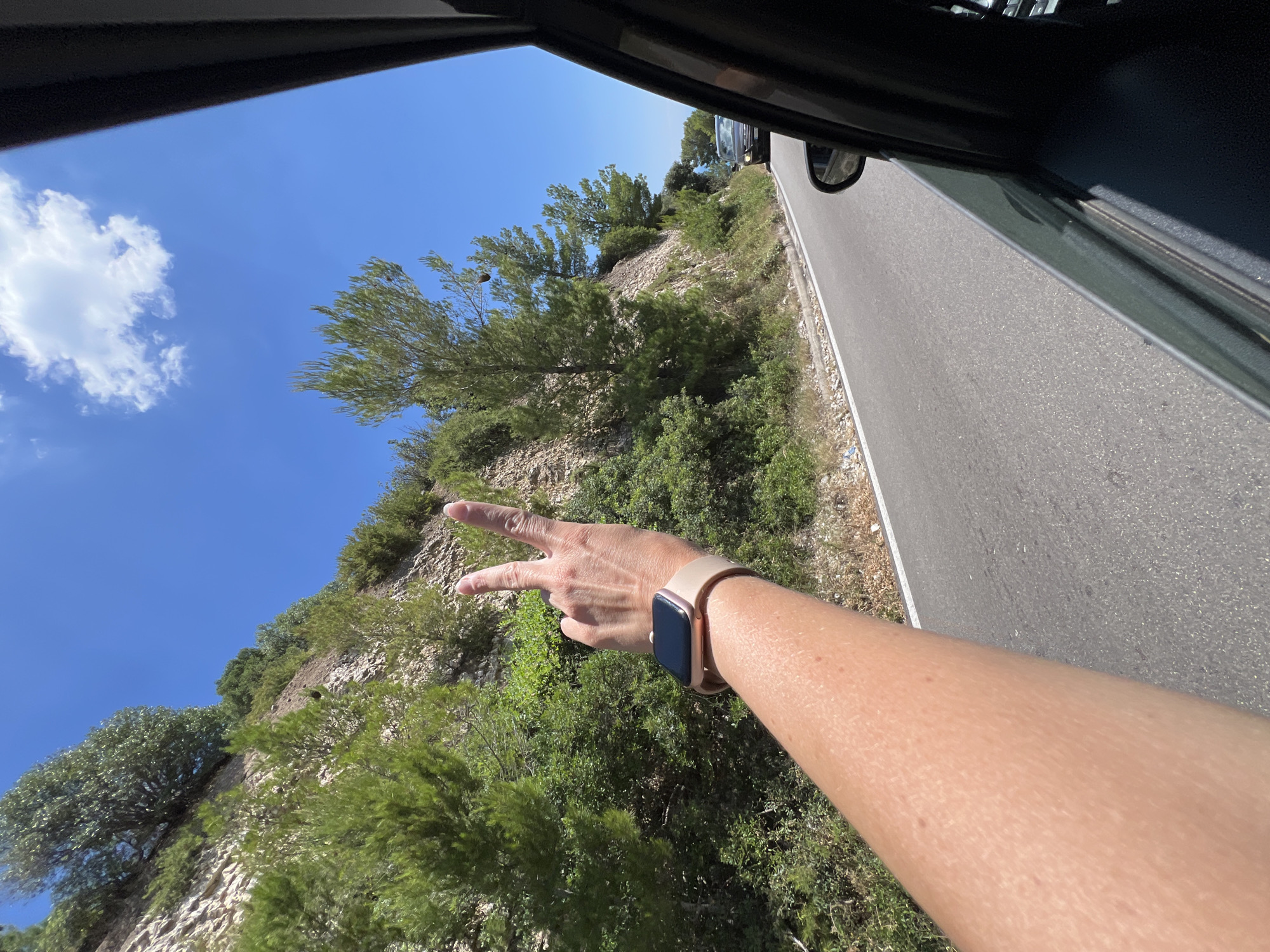 A first-person perspective shot shows a person's arm extended out of a car window, making a peace sign. The arm is tanned and has a light-colored smartwatch on the wrist. The car is driving on a paved road that curves to the right, with a steep, rocky hillside covered in green trees and bushes on the left. The sky is a clear, bright blue with a few white clouds. In the distance, another car is visible on the road. The location is the scenic coastal road near Mattinata, Italy, known for its dramatic cliffs and lush vegetation.