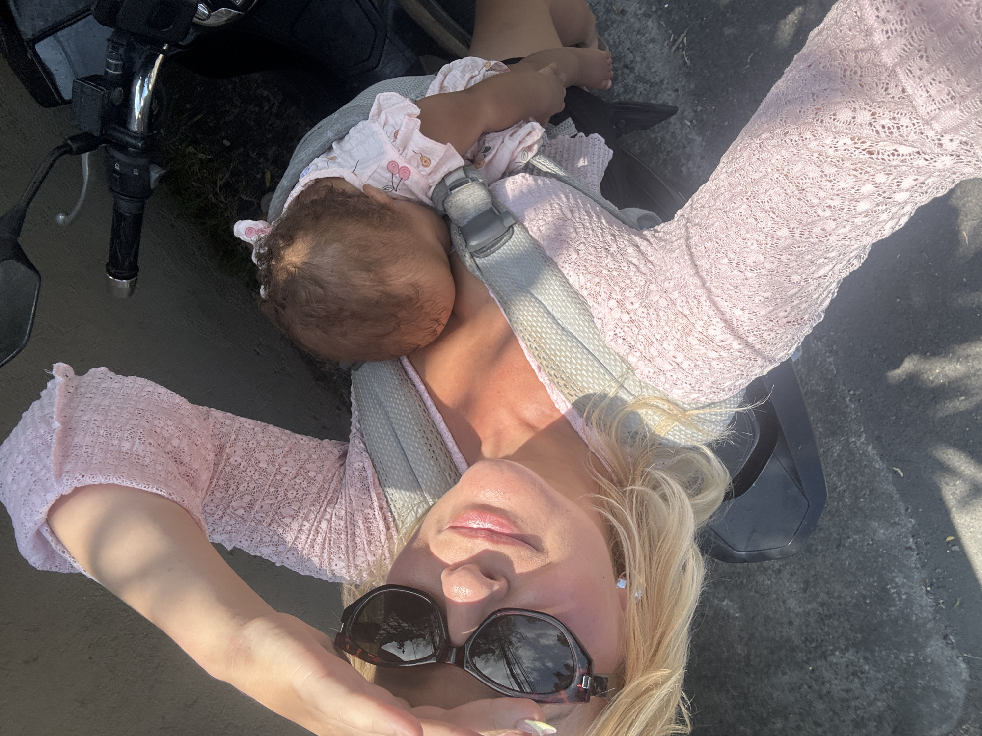 A blonde woman wearing sunglasses and a light pink lace top is carrying a baby in a carrier. She is holding her hand up to shield her eyes from the sun. The baby is asleep. The scene appears to be outdoors, possibly on a street, with a motorcycle visible in the foreground. The ground is paved and appears to be wet in some areas. The overall lighting suggests it is daytime. This image was captured in North Kuta, Indonesia.