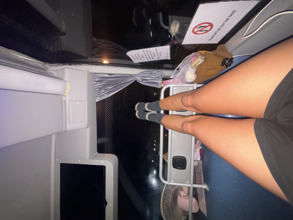 This image depicts the interior of a bus at nighttime, located within Surat Thani City Municipality, Thailand. A first-person perspective captures the lower torso and legs of an individual, clad in black shorts and dark blue socks with light-colored heels. The individual's legs are extended forward, with their feet resting on a silver-colored metal railing or shelf positioned at the front of the bus, near the windshield.
Essential details include:
* **People:** One individual's legs are visible, indicating their presence and action.
* **Actions:** The individual is resting their feet on a designated area. This action occurs directly above a white sign featuring a red circular prohibition symbol with a crossed-out foot, accompanied by the English text: "Please don't put your feet here!"
* **Objects:**
* To the left, on the dashboard, a white portable fan is visible.
* A black speaker is mounted on the metal railing.
* To the right of the railing, a brown bag, partially obscured, holds items contained within a clear plastic bag.
* Blue pleated curtains are tied back on the right side window.
* An overhead rectangular monitor with a dark, unlit screen is visible on the upper left.
* Fluorescent tube lighting illuminates the ceiling.
* A partially visible warning sign on the right window frame advises passengers to "PLEASE KEEP YOUR MONEY AND VALUABLES with you..."
* The bus floor appears to be blue.
* **Environment:** The exterior through the windshield is dark, indicating night, with distant artificial lights suggesting a city environment or transportation area. The interior of the bus is well-lit.
