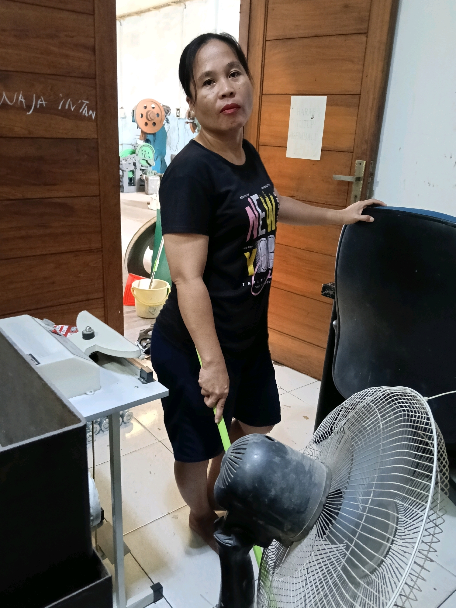 A middle-aged Indonesian woman stands in an indoor workshop, leaning her right hand on a black office chair. She is wearing a black t-shirt with colorful text and dark shorts. In front of her, to the left, is a white tabletop machine with various attachments. A desk fan is visible in the foreground, partially obscuring the lower half of the woman and the machine. The background shows wooden doors, industrial equipment, and a yellow bucket. The scene appears to be in a workshop located in Belimbing, Indonesia, indicated by the text on the wooden door and the presence of industrial machinery.