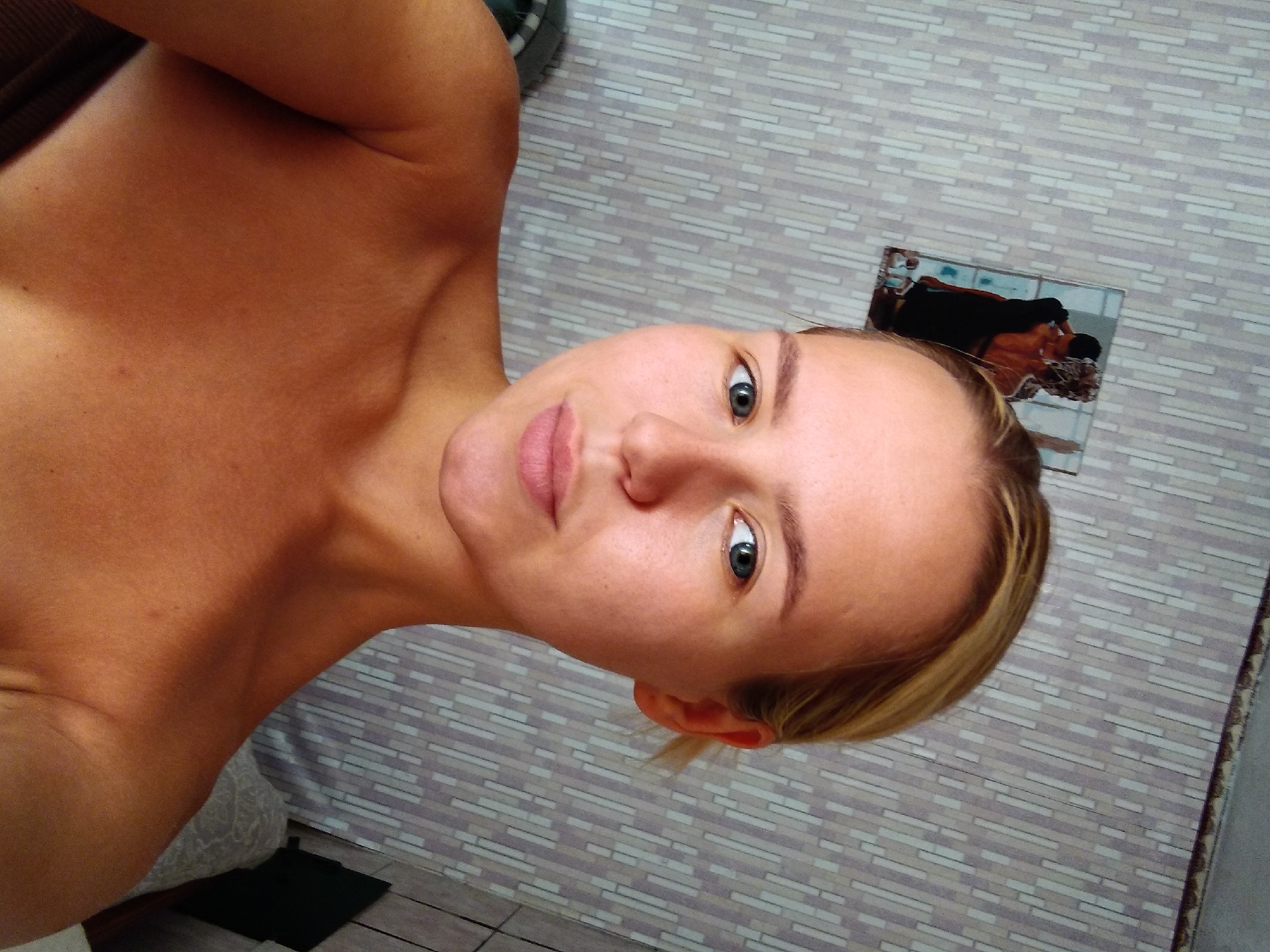 The media file is a close-up, portrait-style photograph of a young woman. She is facing the camera, with her head tilted to the side and slightly upwards. Her eyes are open and looking directly at the viewer, and she has a neutral or slightly amused expression. Her lips are parted slightly. She appears to have fair skin with a warm undertone and light brown hair. She is topless, with her left shoulder and upper chest visible. The background is a wall with a textured pattern resembling light-colored bricks or tiles. A small framed photograph is attached to the wall in the upper right portion of the image, depicting two people embracing. The lighting suggests it is daytime, likely indoors, given the wall texture and the artificial light source that creates subtle shadows. The overall mood is casual and personal.