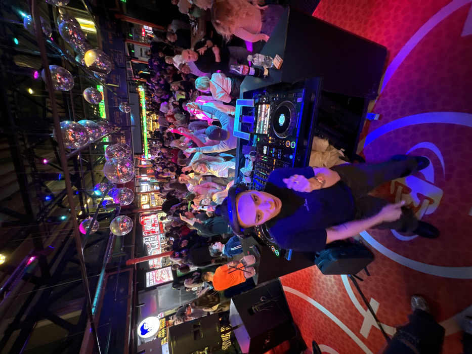 The image depicts a crowded, energetic nightclub scene within Depo Moscow (Депо Лесная), a large food market and entertainment complex located at Lesnaya Ulitsa, 20, building 3, Moscow, Russia.
The scene is set indoors, characterized by high ceilings with exposed industrial-style beams and ductwork. Numerous small, reflective disco balls hang from the ceiling, reflecting a kaleidoscope of multicolored lights, predominantly blues, purples, and pinks. The overall lighting is dim, augmented by vibrant neon and LED strip lights in green, red, and blue outlining various bars and stalls in the background. The floor in the foreground and central area is distinctive, featuring a red surface with white markings resembling a stylized basketball court.
In the immediate foreground, a female DJ, appearing to be in her late 20s to early 30s, is actively performing. She wears large black over-ear headphones, a dark blue short-sleeved t-shirt with a small white logo, and dark trousers. Her left arm is outstretched, and her mouth is open, suggesting she is engaging with the crowd, speaking, or singing. She stands behind a black professional DJ console equipped with two players and a central mixer. On the DJ booth, to the left, are two pink cocktails in glasses with straws, and to the right, a Coca-Cola bottle, a plastic water bottle, and a pack of cigarettes. Large black speakers are positioned to the left of the DJ booth.
Behind the DJ, a dense crowd of approximately 100-150 individuals, primarily young adults, fills the central and mid-ground. Many are actively dancing, raising their hands in the air, or looking towards the DJ, indicating an energetic and engaged atmosphere. The crowd exhibits casual club attire. In the far background, along the perimeter of the venue, various illuminated signs with Cyrillic text are visible, indicating different food and drink vendors, consistent with a food hall environment. One sign on the far left features a blue cartoon character, strongly suggesting a "Kroshka Kartoshka" (Russian baked potato chain) stall within the complex.
The image is captured from a slightly elevated, wide-angle perspective, looking down towards the DJ and across the animated crowd, creating an immersive view of the lively event.