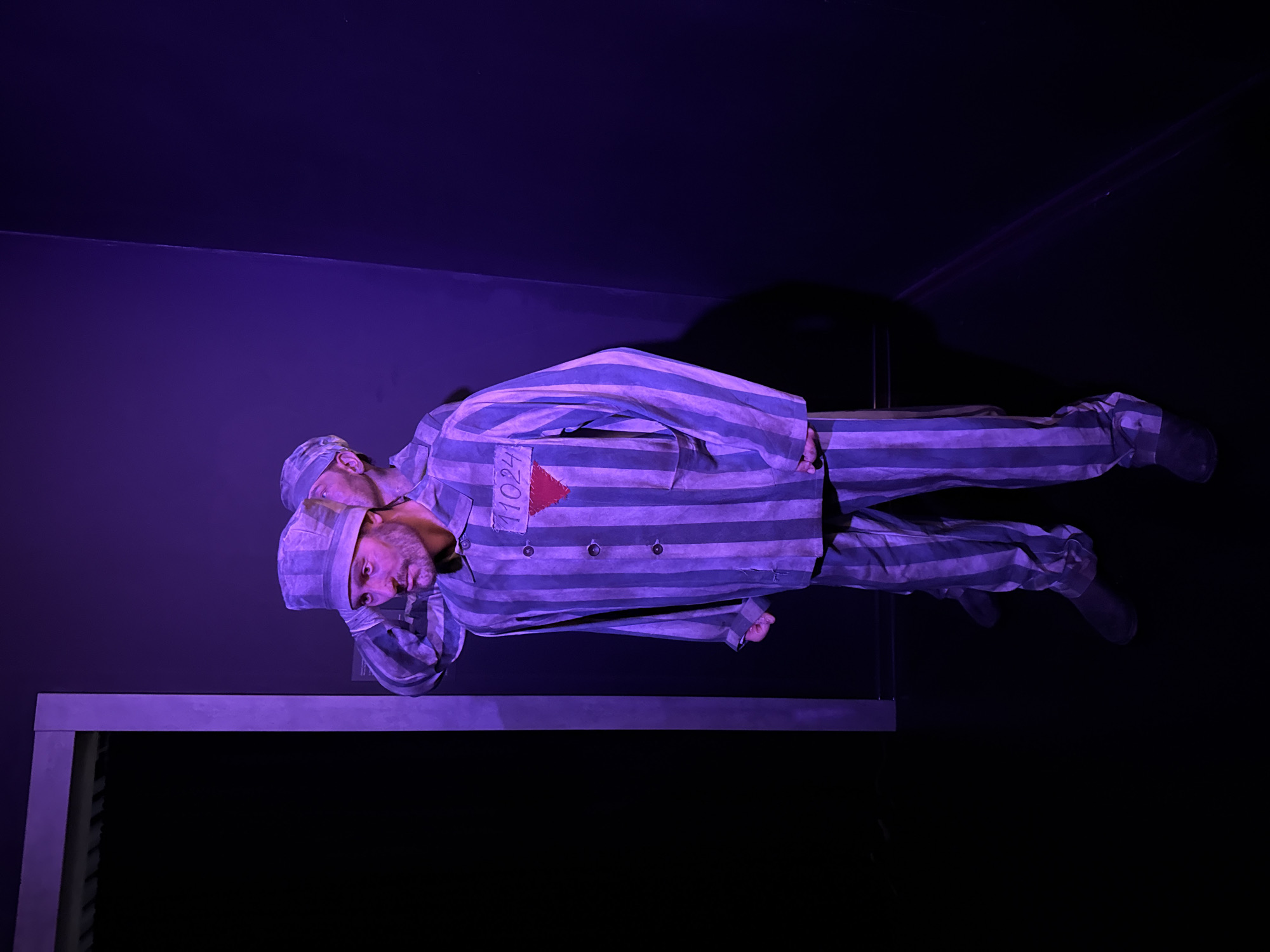 This is a photograph of two people on a stage, illuminated by purple light against a dark background. The two individuals are dressed in striped prison uniforms, consisting of a jacket and pants, and head coverings. One person is positioned in the foreground, lying on their back, with their head slightly raised. They have a beard and appear to be looking upwards. The other person is behind and above the first, their body also clad in a striped uniform, with their head resting on the chest of the person in front.
A prominent detail on the jacket of the person in the foreground is a red triangle with the number "11024" written in white. The clothing is loose-fitting and appears to be made of a coarse fabric. The setting suggests a theatrical performance or reenactment, possibly depicting a historical event or a scene of imprisonment. The lighting creates a dramatic and somber mood. The backdrop is dark, with some abstract light patterns visible in the upper right portion of the image, possibly from stage lighting. There are no obvious cues regarding time of day or weather, as the scene is indoors and artificially lit. The emotion conveyed is one of solemnity and perhaps despair. The visible text is "11024".