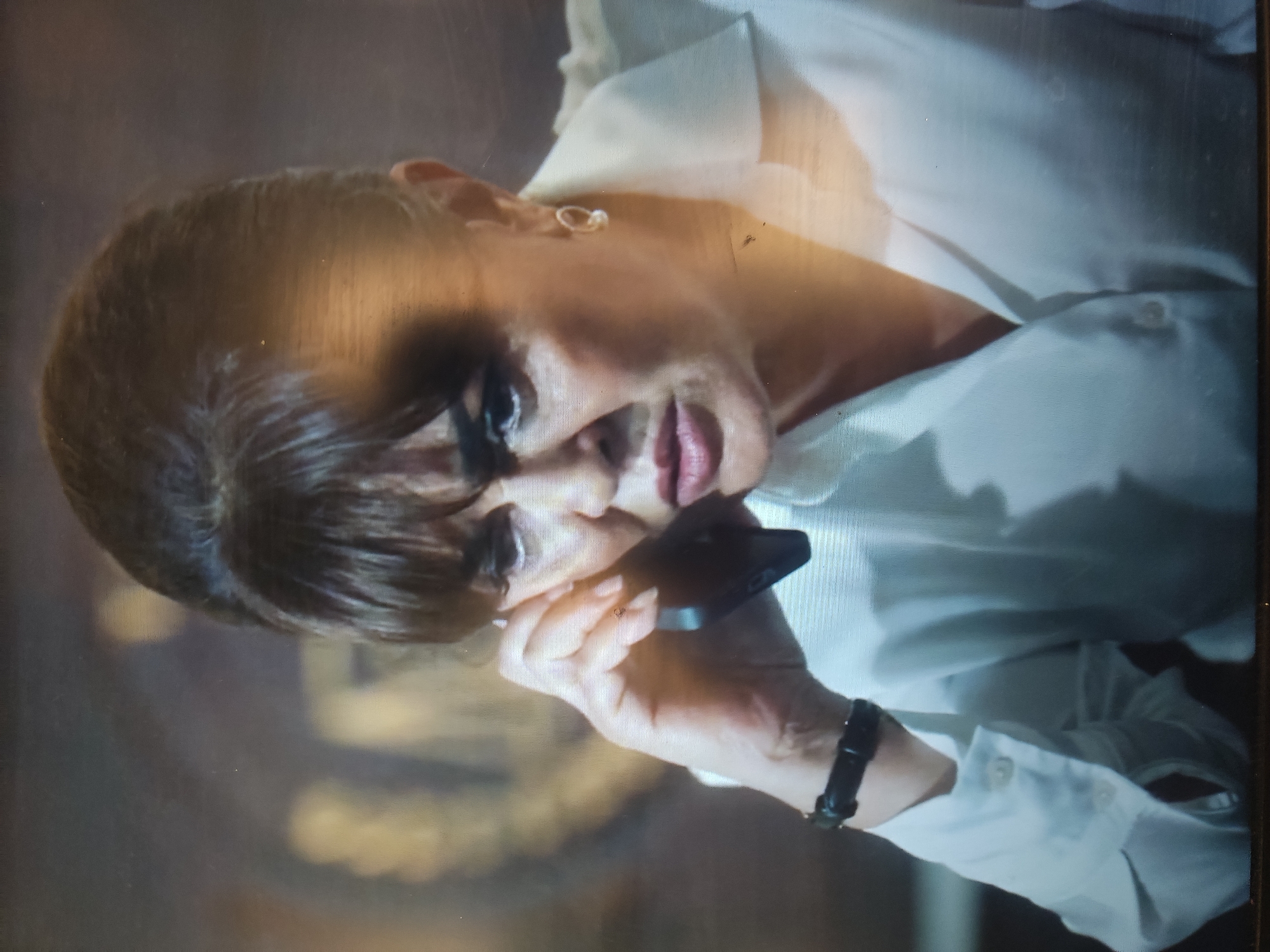 The image depicts a close-up, horizontally oriented shot of a woman with dark hair, wearing a white collared shirt. She is holding a black object, possibly a mobile phone, to her ear, suggesting she is engaged in a phone conversation. Her expression appears serious or contemplative, with her eyes looking downwards and slightly to the side. A subtle gold earring is visible in her left ear, and a black bracelet adorns her right wrist. The lighting creates a dramatic effect, with highlights and shadows on her face and clothing. The background is out of focus, with muted, dark tones, offering no specific environmental cues. The overall impression is one of intimacy and focus on the woman's private moment. There is no visible text in the image. Given the provided location context of Klyuchi, Russia, the scene could be indoors, during the daytime or evening, as there are no clear indicators of weather or time of day.