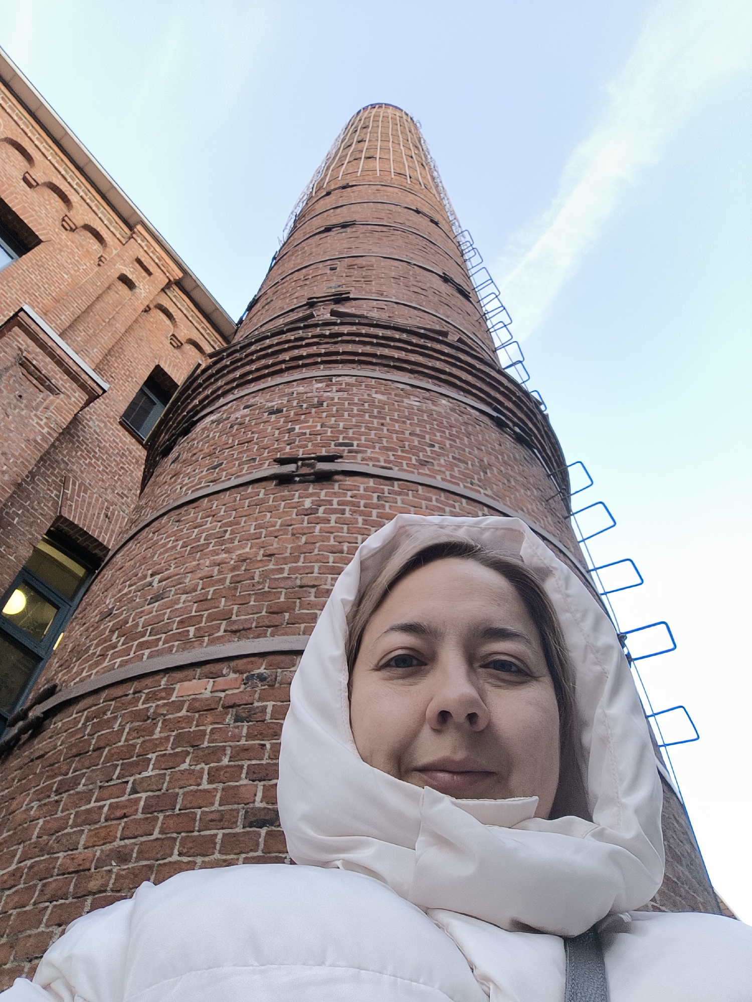 A woman takes a low-angle selfie, capturing herself against the backdrop of a tall brick chimney and an adjacent brick building, likely in Moscow, Russia. The woman, positioned in the lower center of the frame, looks directly at the camera with a neutral to slight smile. She wears a white or light cream hooded puffer jacket, with the hood up, framing her face.
The dominant feature in the background is a towering, cylindrical industrial chimney made of red brick, extending upwards from behind the woman's head and filling much of the right side of the frame. It features horizontal metal bands at intervals and a blue, caged ladder running vertically up its side. To the left of the chimney, a multi-story brick building with distinct arched architectural details and windows is visible. The buildings share a similar red brick construction, suggesting a cohesive architectural style, possibly from a former industrial complex.
The sky above is bright and clear with a light blue hue, indicating daytime. A faint contrail is visible in the upper right portion of the sky. The extreme low-angle perspective exaggerates the height and imposing nature of the chimney and building. There is no visible text in the image.