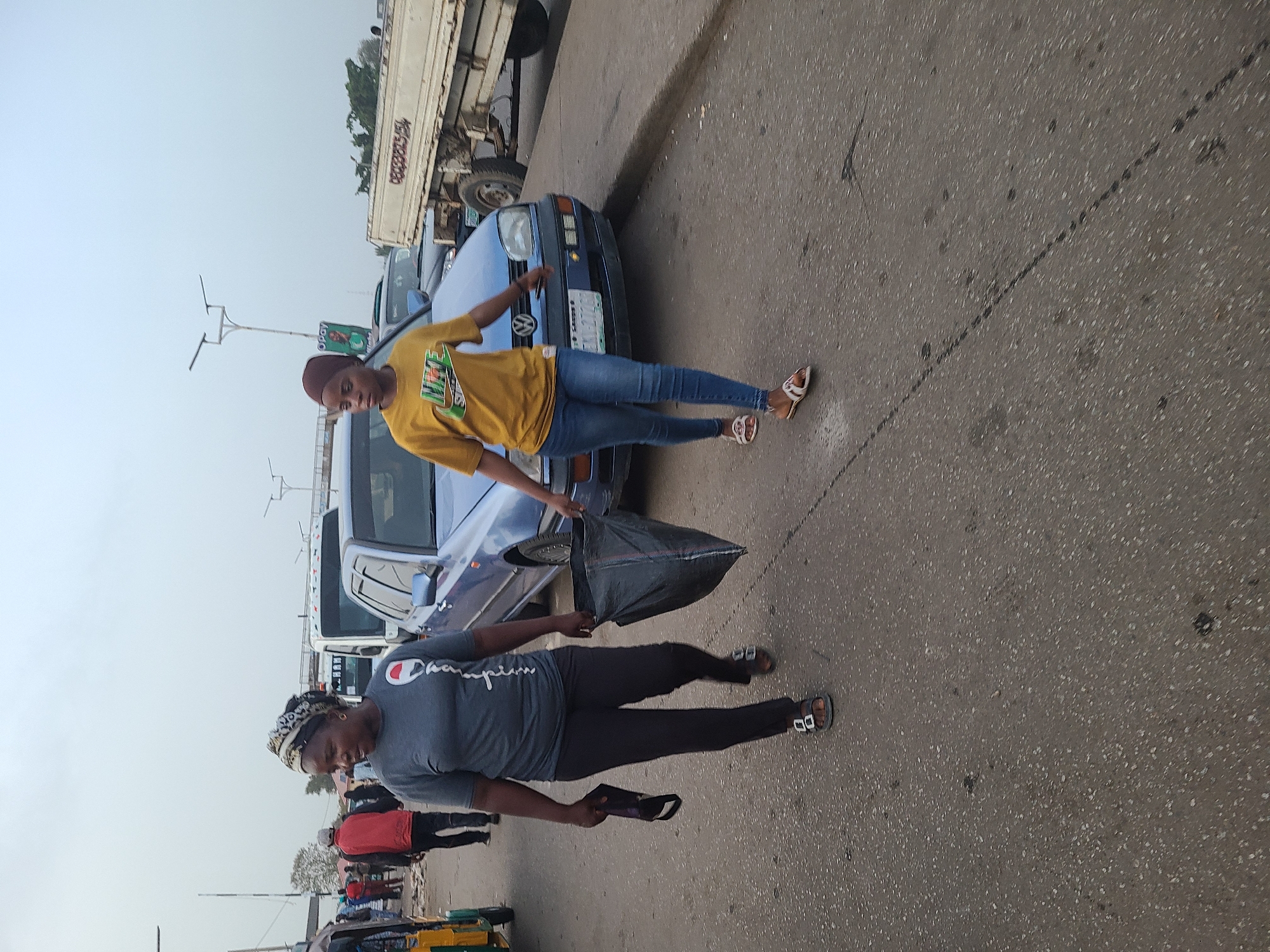 Two women stand on a grey asphalt road next to several parked vehicles, including a blue Volkswagen and a large white truck. The woman in the foreground wears a grey Champion T-shirt and black pants and is carrying a black bag and a black face mask. The second woman wears a yellow T-shirt with a green design and blue jeans. The scene includes street lights and other pedestrians in the background. The location is Jos, Nigeria.