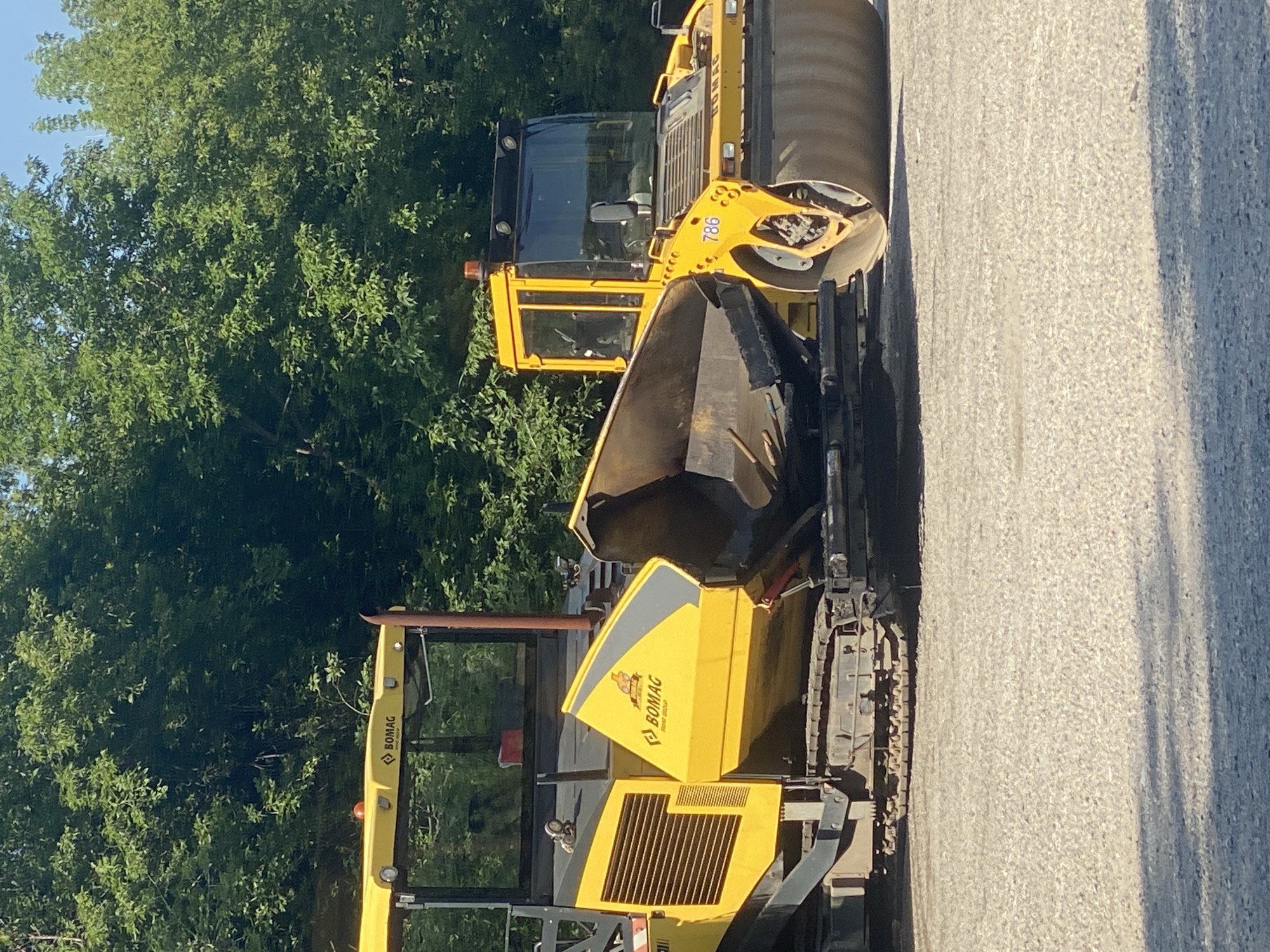 In Novokuznetsk, Russia, a yellow BOMAG asphalt paver and a yellow BOMAG tandem roller are positioned on a gravel surface adjacent to a line of trees. The paver is equipped with a hopper and track system, while the roller features a large drum and cab. The scene is illuminated by daylight, casting shadows on the ground.