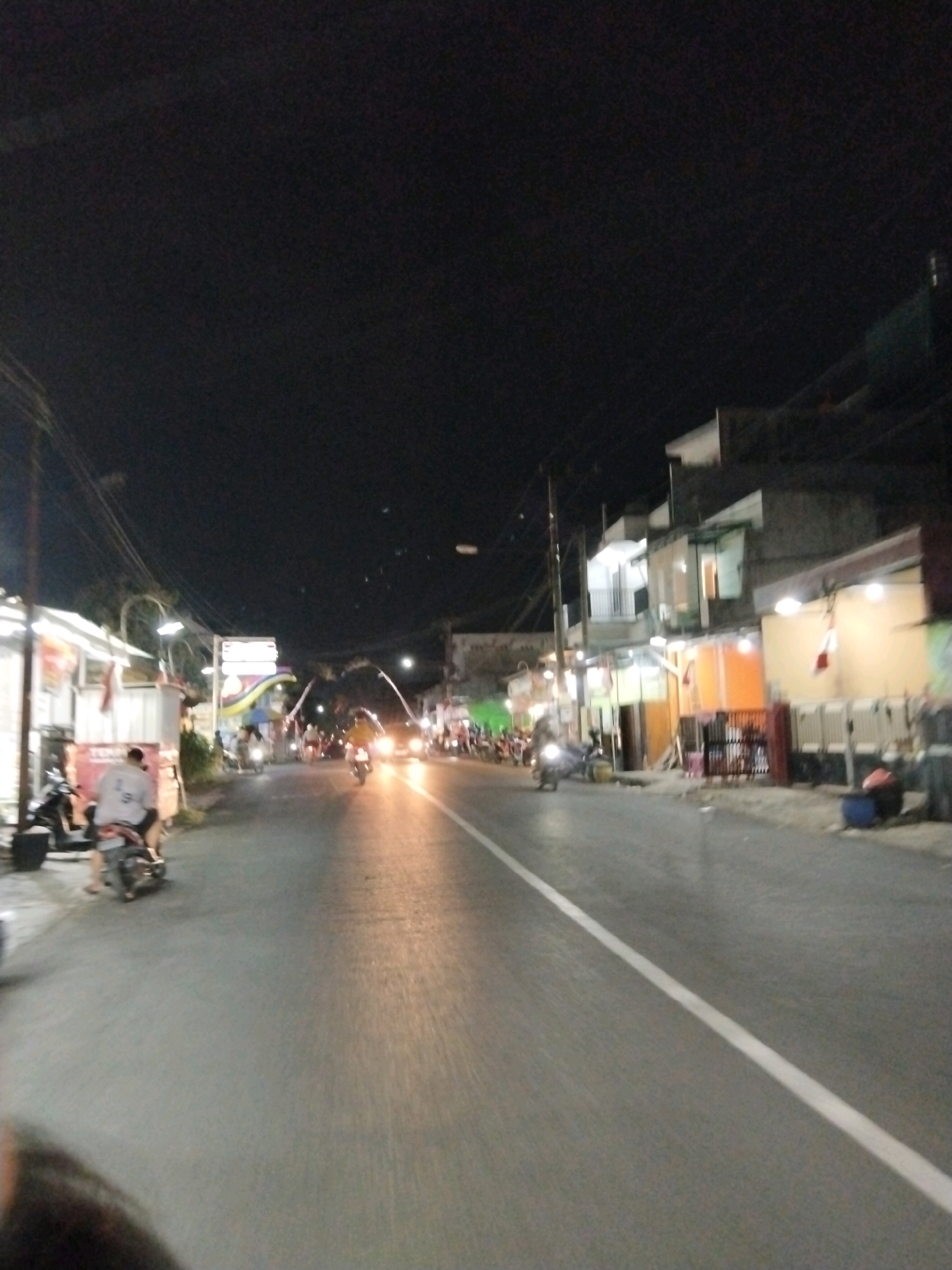 The image captures a nighttime scene on a street in Banjararum, Indonesia. The road is paved with asphalt and has a white line marking its center. On the left side of the road, there are low-rise buildings with illuminated storefronts, a motorcycle parked near a building, and a person on a motorcycle in the foreground. Further down the street, more motorcycles are visible, some with headlights on, indicating movement. The right side of the road features multi-story buildings, some with illuminated windows and balconies. Streetlights are present, casting a yellowish glow on the road. The sky is dark, typical of nighttime. The overall impression is of a moderately busy street in a town or city at night.