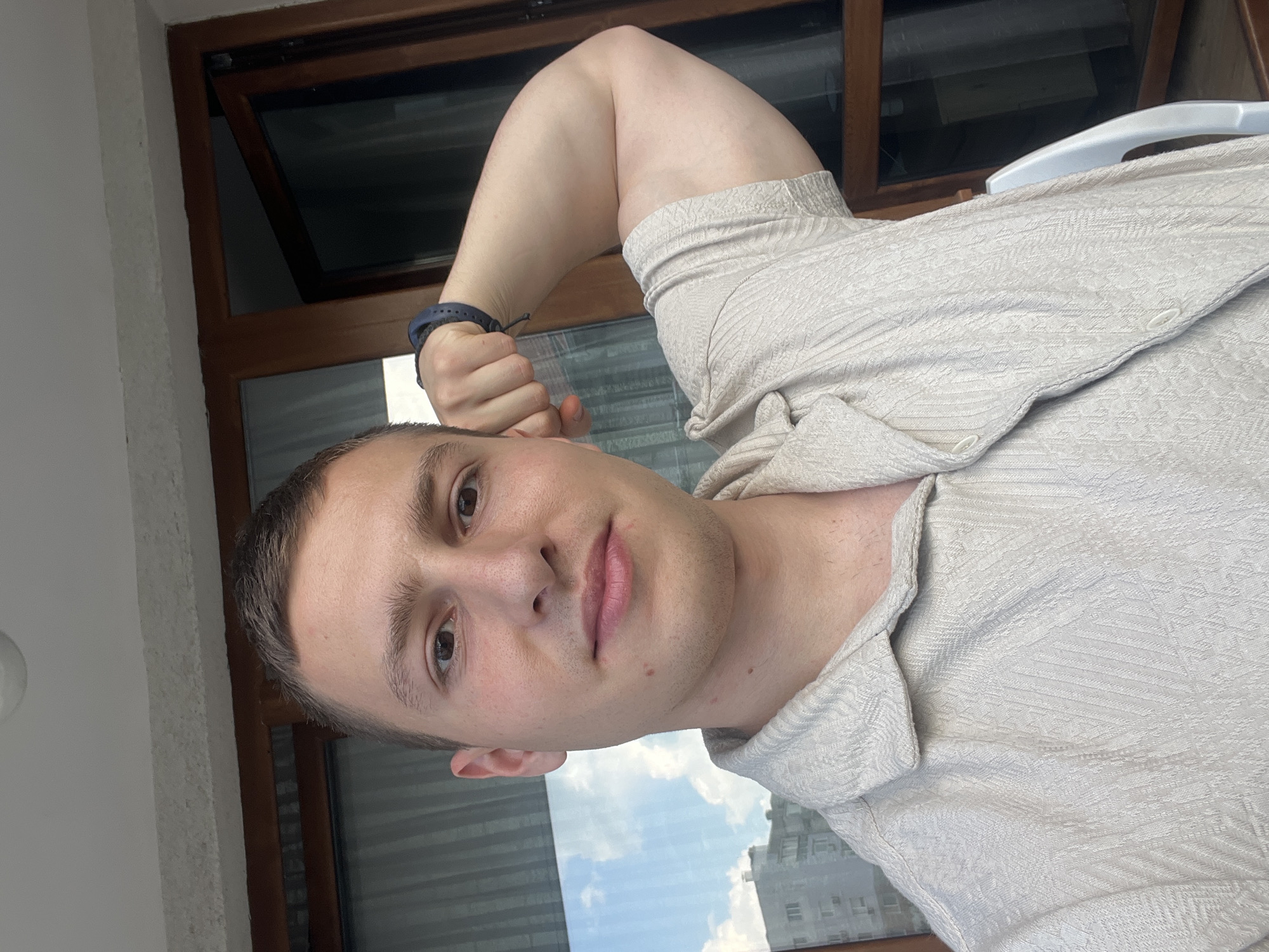 The image shows a man posing in what appears to be an apartment or balcony. The location is indicated by the view through the window as being in Varna, Bulgaria.
The man is fair-skinned with short brown hair and brown eyes. He is wearing a light-colored, button-down shirt. He is flexing his right arm, which is bent at the elbow and positioned near his head. He is also wearing a dark blue bracelet on the wrist of the same arm. His facial expression is neutral.
Behind the man is a window with a brown wooden frame. Through the window, one can see a cityscape, including buildings and a blue sky with clouds. A glimpse of a white plastic chair is also visible. The background also suggests a view of the sky and potentially buildings, further reinforcing the location within a city. The photograph has been rotated and shot indoors in daylight.