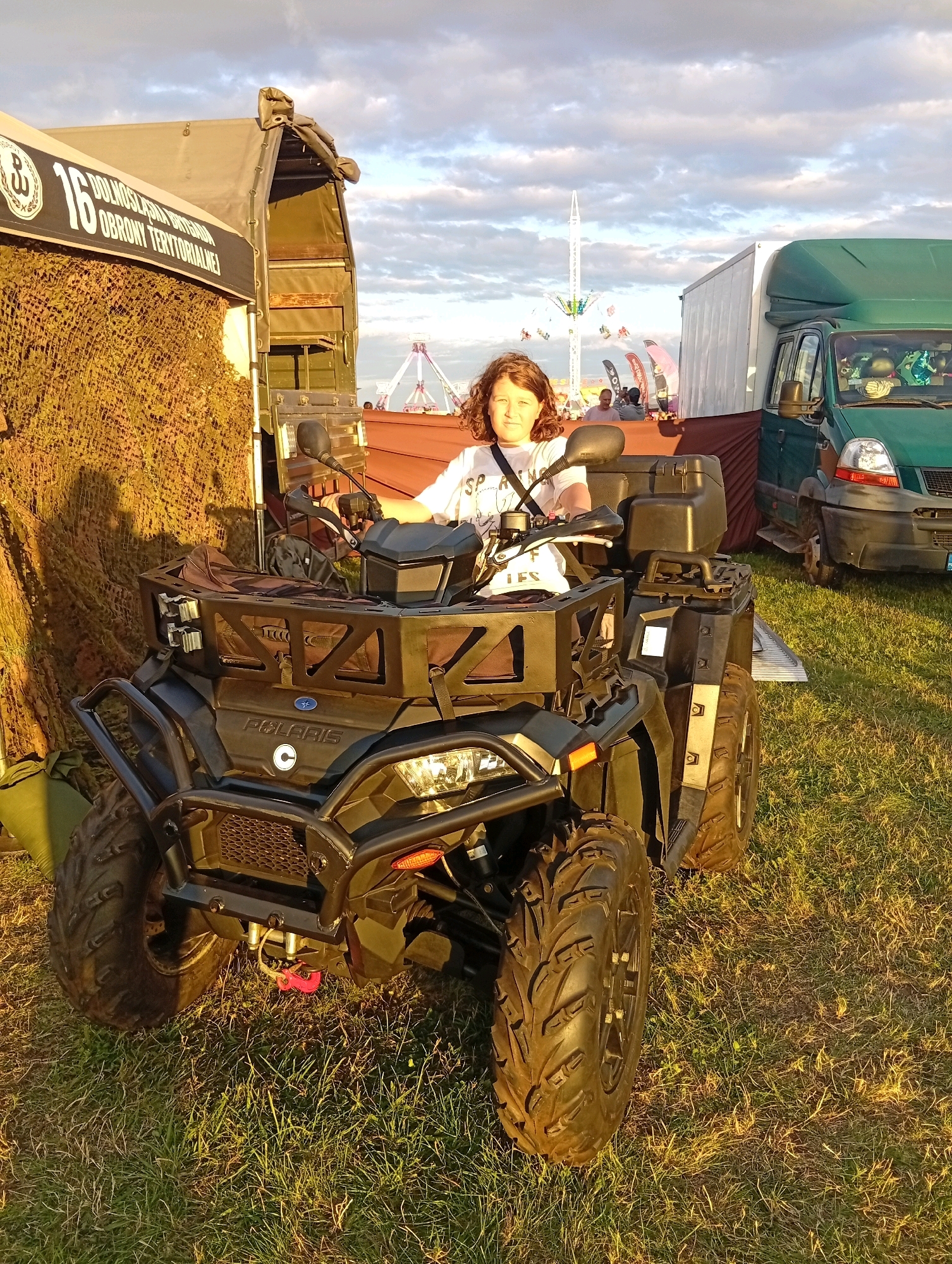 A young person with curly brown hair, wearing a white t-shirt with black lettering, sits on a black Polaris ATV. The ATV is positioned in a grassy area, with a camouflage net draped over a structure to its left. Behind the ATV, there is a large tent with text that includes "16 DOLNOŚLĄSKA OBRONY TERYTORIALNEJ". Further in the background, a green truck is parked, and a fairground ride with colorful lights is visible. The sky is partly cloudy, and the scene is illuminated by the warm light of what appears to be late afternoon or early evening. The image was taken in Szewce, Poland.