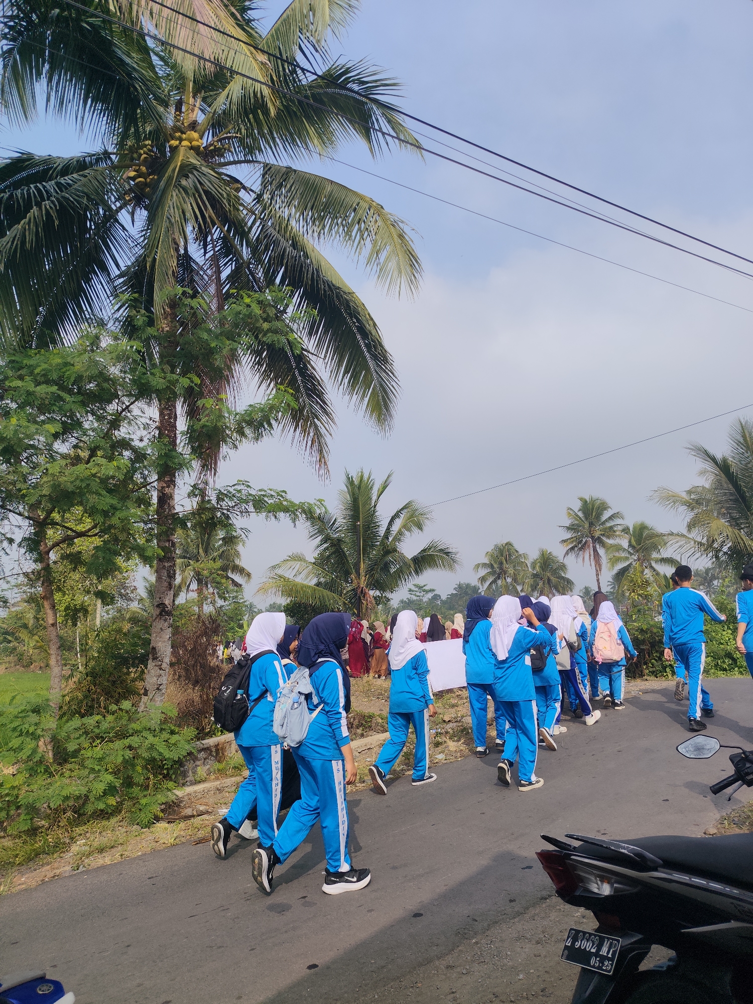 In Sukaratu, Indonesia, a group of schoolchildren in blue uniforms and hijabs are walking along a paved road lined with palm trees. The sky is cloudy, and the weather appears to be warm. The children are carrying backpacks and some are holding a large white banner. The road is empty except for the children and a motorcycle parked on the side of the road. The scene suggests a school event or a parade.