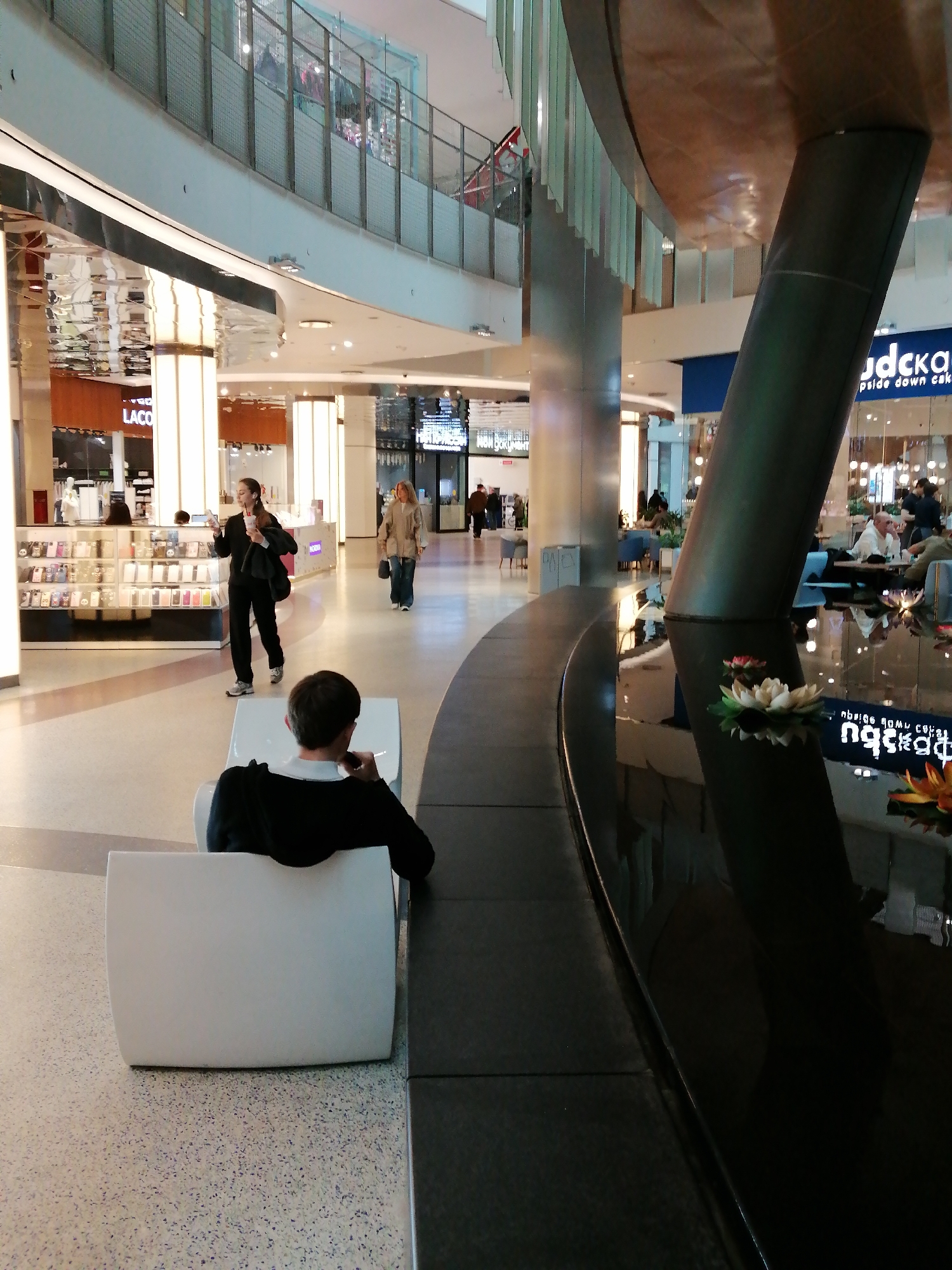 The image depicts an interior scene of a commercial space. A person sits on a white chair in the foreground, facing away from the viewer. A woman in dark clothing walks while holding a beverage. Another woman walks further away. The space appears to be a shopping mall, with various retail stores visible, including a Lacoste store. The interior features a water installation with flowers, near a support structure. The reflected text "ПЯСКА" is visible in the water. The scene takes place in the Evropeisky Shopping Mall, a Moscow location.