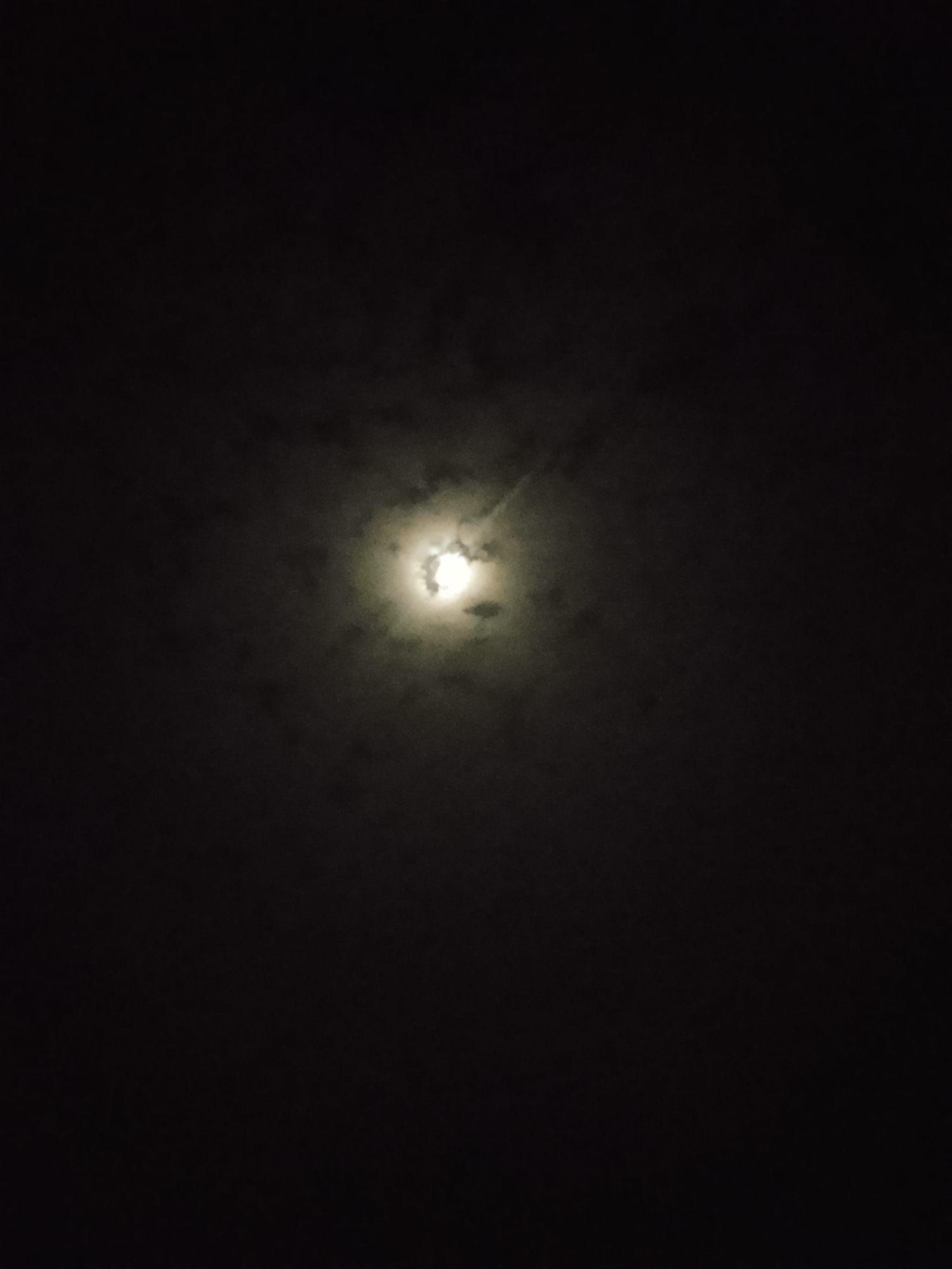 The image depicts a dimly lit night sky with a bright moon partially obscured by clouds. The moon is located in the upper center of the frame and appears to be full or nearly full. The surrounding sky is dark, with wispy clouds creating a textured effect around the moon. There are no visible landmarks or features that would indicate the specific location within the city of Kerang, Nigeria. The scene is devoid of any people, animals, or man-made objects, focusing solely on the celestial phenomenon.
