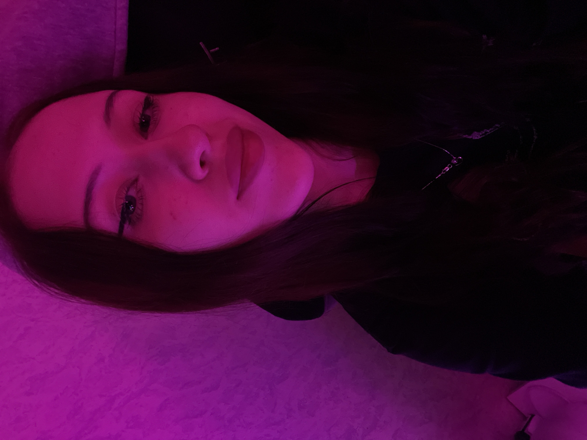The image shows a close-up, selfie-style shot of a young woman's face and upper body, illuminated by pink lighting. She has long, dark hair, styled down. Her makeup includes winged eyeliner and mascara. She is wearing a dark-colored top, possibly a sweatshirt or t-shirt, and a delicate necklace with a small pendant. The background is a textured wall, also lit with pink light, with a darker area to the right of the subject. The overall impression is of a candid, indoor shot with dramatic lighting. The location in Votkinsk, Russia, is not visually discernible from the image itself.