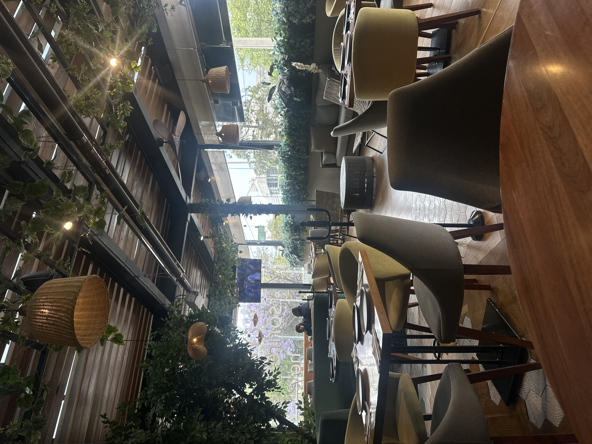 The image shows the interior of a restaurant, presumably in Huixquilucan de Degollado, Mexico. Rows of dining tables with upholstered chairs are aligned along a long axis. The chairs are primarily a muted green hue with light wood frames. The tables appear to have dark green surfaces. Floor material changes along the length of the room from tiles to wooden planks. Large windows, visible in the background, are partially obscured by dense greenery, offering a glimpse of a cityscape outside. There is a TV monitor along the window. Wicker-style lamps are suspended from the ceiling amidst decorative foliage. The interior design incorporates wooden planks, metal beams, and suspended greenery, creating a garden-like atmosphere. Some people sitting near the window are visible, but they are far away.