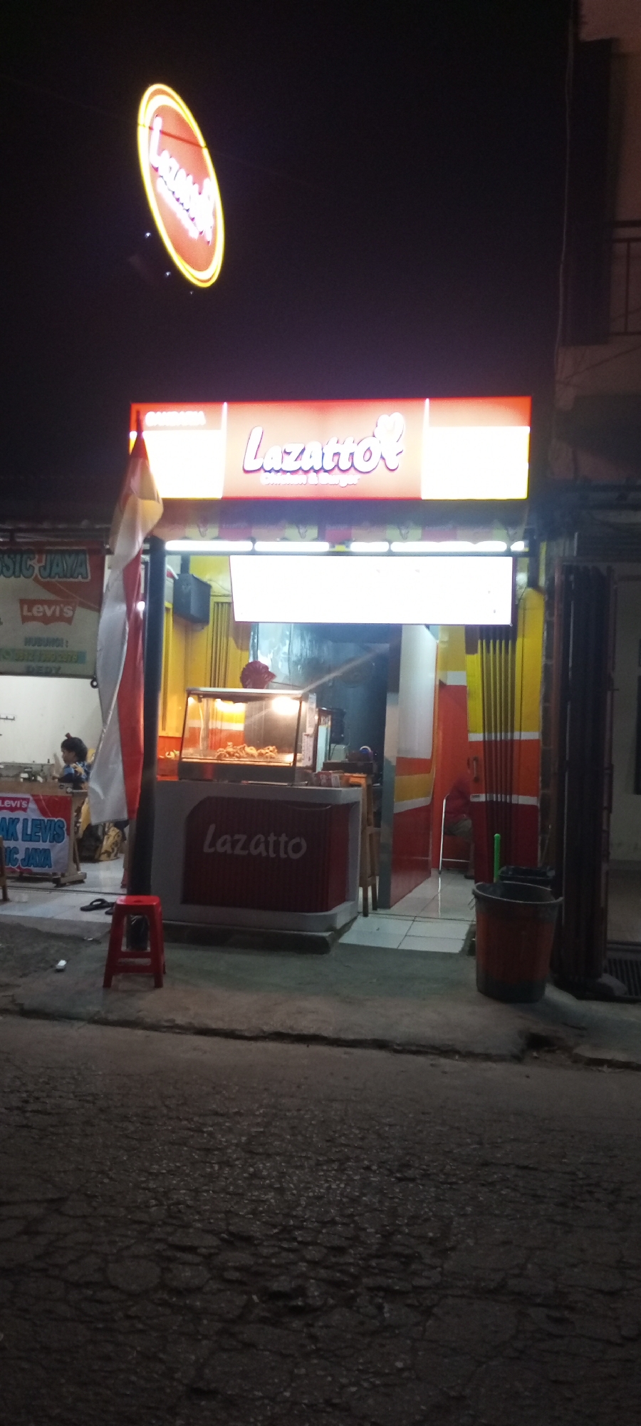 The image shows a storefront at night, likely in Setu, Indonesia. A prominent sign displays the name "Lazatto" in stylized pink lettering, with "Chicken & Burger" below it. The sign is illuminated with yellow and red lights. To the left of the main sign, a circular illuminated sign also reads "Lazatto." The storefront itself is painted in yellow and orange vertical stripes. In front of the store is a display case filled with fried food, and a counter with a red base also has "Lazatto" written on it in white letters. A small red stool is placed on the pavement in front of the counter. To the left of the storefront, another sign is visible with text that appears to be "MUSIK JAYA" and "Levi's". In the background, there are some vertical poles and a glimpse of a building. The ground in front of the storefront is asphalt, showing signs of wear and cracking. A dark-colored bucket with a green handle sits near the right edge of the frame. A person can be seen through the display case, possibly an employee. The overall scene depicts a food stall or small restaurant operating at night.