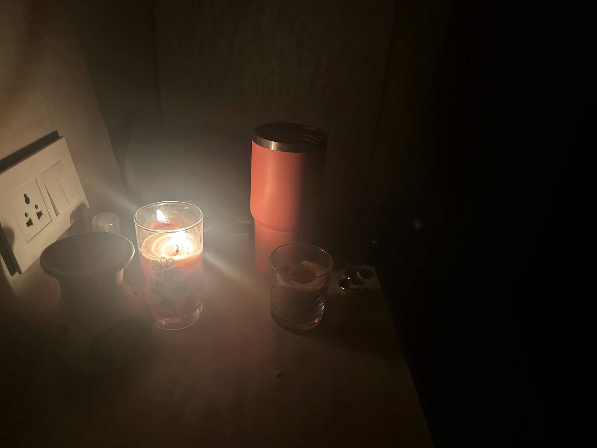 The image depicts a dimly lit scene on a wooden surface, likely a table, within an indoor environment. The lighting is predominantly from a lit candle in a glass, casting a warm glow on its immediate surroundings. To the left of the candle is a white electrical socket with two switches. To the right of the candle, there is a small glass with some liquid, and further to the right, a tall, orange insulated tumbler. In the background, the scene fades into darkness, with a hint of a wall or curtain. The overall atmosphere is tranquil and intimate, possibly suggesting a quiet evening or a moment of reflection. The setting, as per the prompt, is within the city of Lalitpur, Nepal.