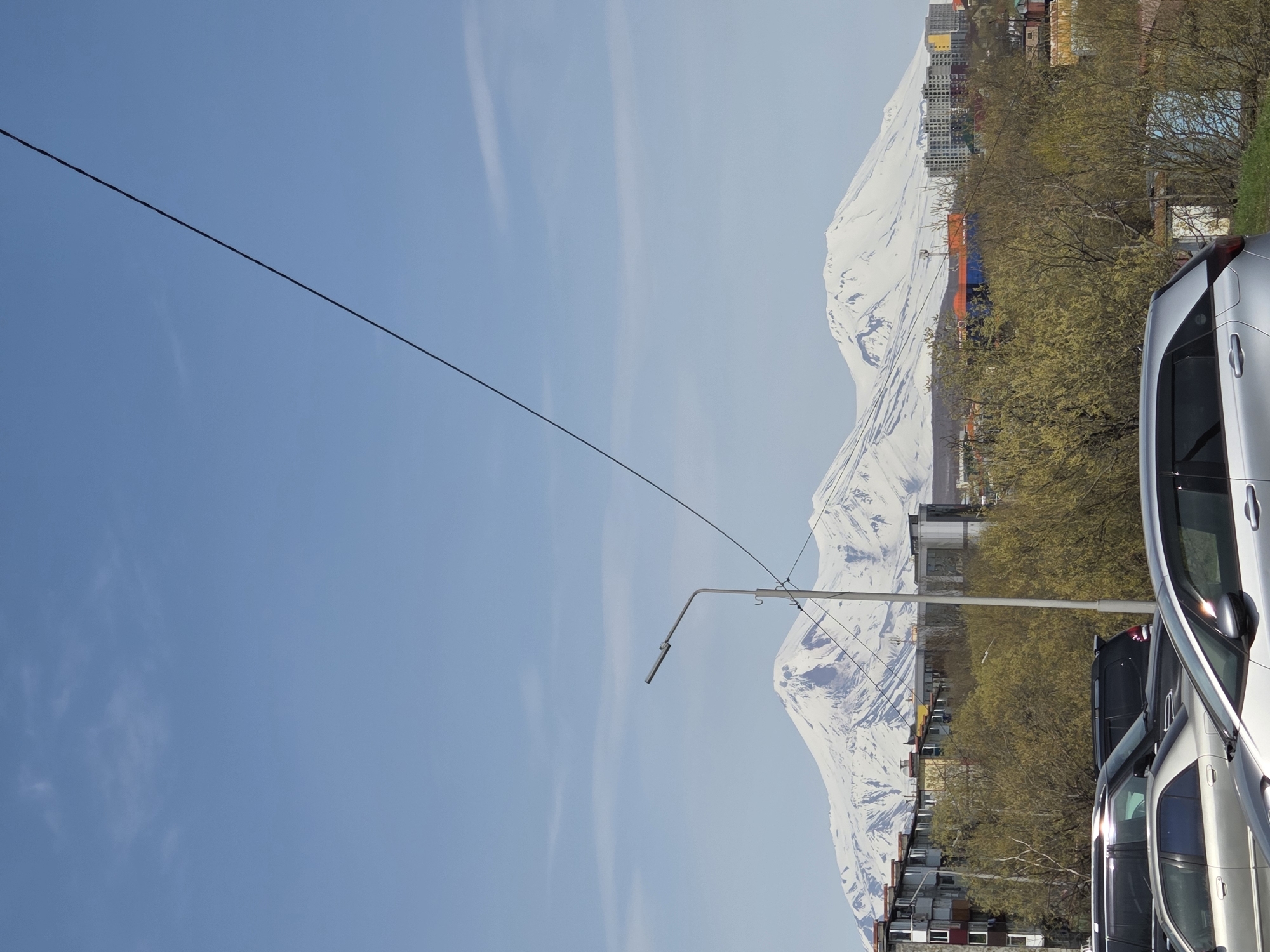 The image captures a scene in what appears to be an urban setting with a snow-capped mountain in the background. The sky is predominantly blue with light clouds. A power line stretches across the upper left portion of the image. In the middle ground, there is a street lamp and various buildings. A row of parked cars is visible in the foreground.