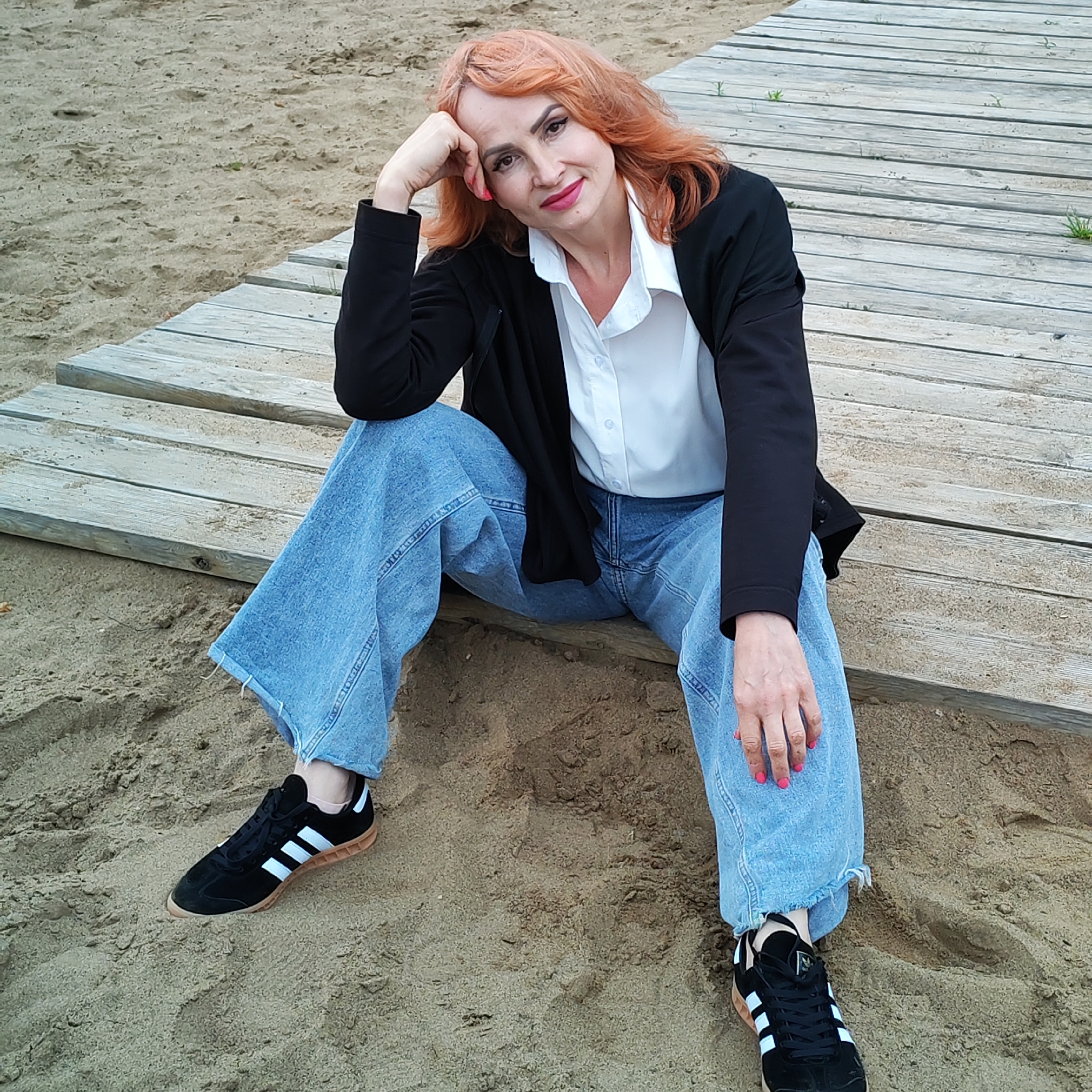 An individual is depicted sitting outdoors on a sandy surface in Chaikovsky, Russia, adjacent to a wooden walkway. The person is a middle-aged female with wavy, reddish-orange hair, facing forward with a slight smile and wearing visible makeup including dark eyeliner and pink lipstick. She is dressed in a white collared shirt, an unbuttoned black blazer, and light blue, wide-leg denim jeans with frayed hems. Her footwear consists of black Adidas sneakers with white stripes and a light brown gum sole. Her left hand is positioned against her temple, and her right hand rests on her right thigh; both hands exhibit pink nail polish. The ground is composed of light brown sand, and a light-colored wooden plank path extends diagonally through the scene, with sparse green vegetation visible between the planks and in the sand.