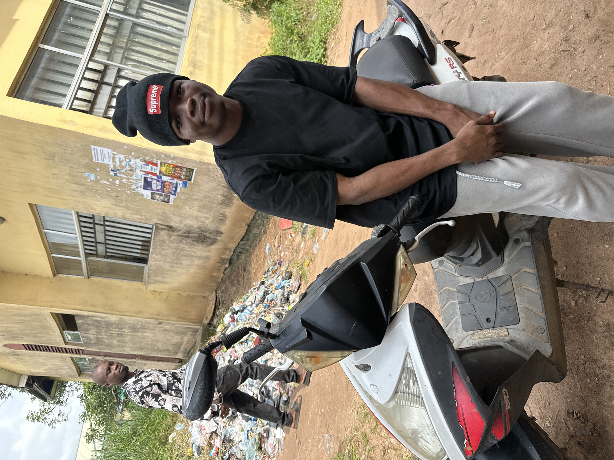 In Ihiagwa, Nigeria, a young man wearing a black Supreme beanie, a black t-shirt, and grey sweatpants is seated on a parked scooter. He is looking at the camera with his hands clasped in front of him. Behind him, another man in a patterned shirt walks away from the camera, his back to the viewer. The scooter is parked on a dirt road next to a pile of trash. A yellow building with barred windows is visible in the background, with posters taped to its exterior. The scene appears to be in an outdoor, urban setting.