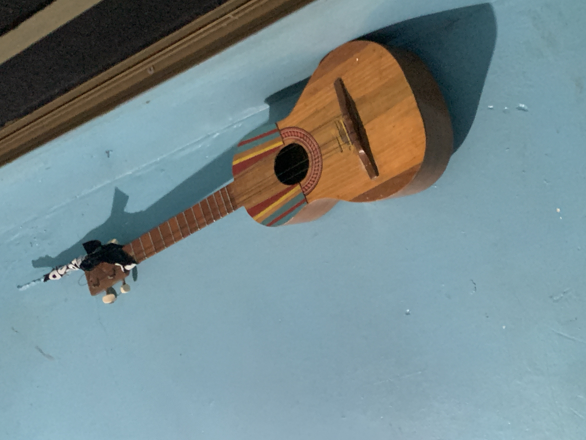 A small, wooden stringed instrument, possibly a ukulele or a small guitar, is hanging on a light blue wall. The instrument has a natural wood finish and features colorful stripes of red, yellow, and blue above the soundhole. The soundhole itself is dark and circular, with a decorative red and yellow border. The bridge is visible on the body of the instrument, and strings are present. The headstock has tuning pegs and is adorned with a black and white patterned fabric wrap. The background is a textured light blue wall, with a shadow cast by the instrument to its right. The upper right of the image shows a glimpse of a dark surface and a wooden frame, possibly a doorway or window. The image appears to be taken indoors, in a residential or casual setting. There is no indication of Santa Ana, Venezuela, in the visual elements of the image.