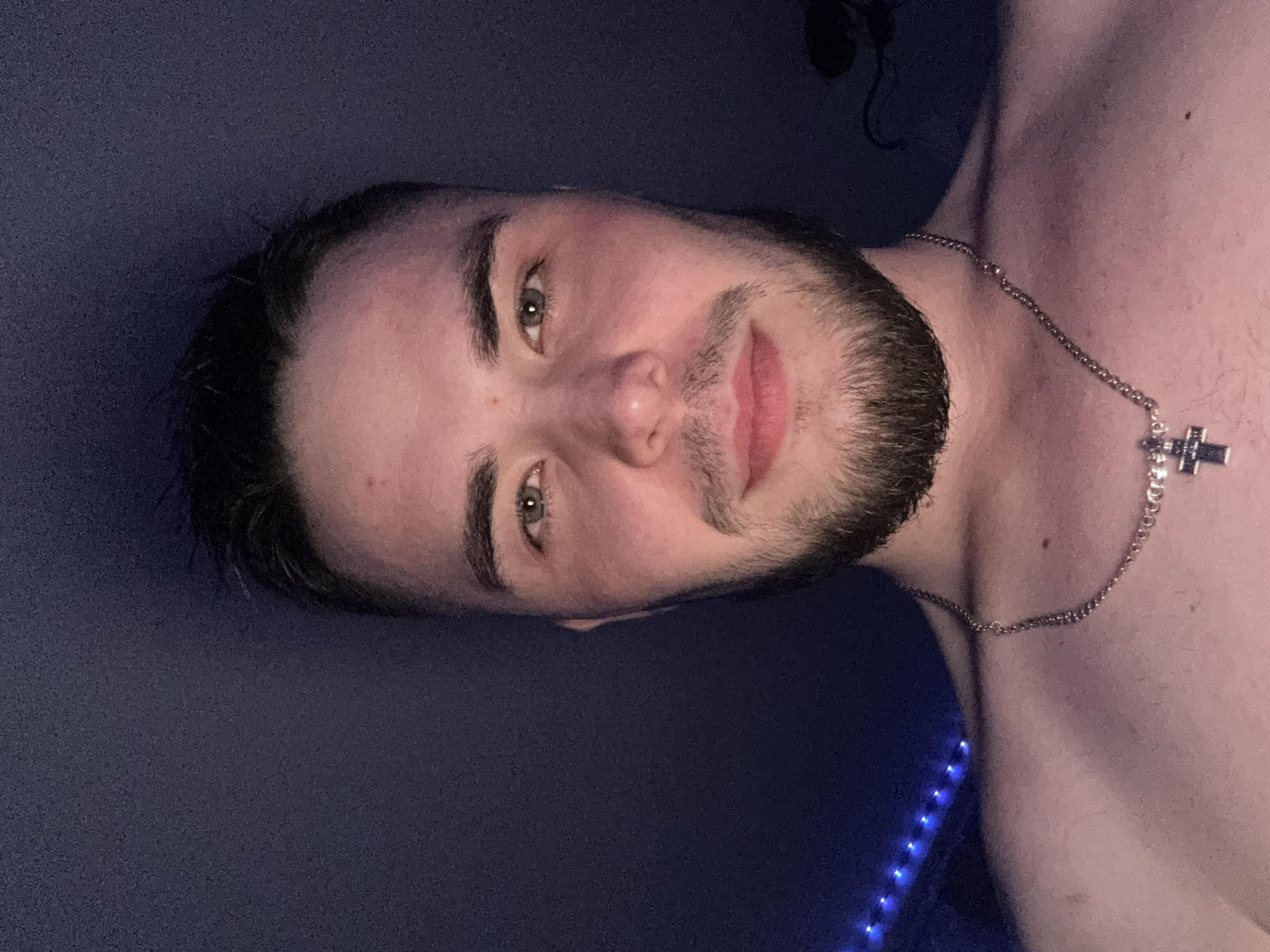 A close-up portrait shows a young man with light skin, dark hair, green eyes, and a dark, short beard or heavy stubble. The image is rotated 90 degrees clockwise. He has full lips and looks directly at the camera with a neutral to slightly content expression. A silver-colored chain with a prominent cross pendant hangs around his neck. His bare chest and shoulder are visible. The background is a dark, solid color, possibly a wall, with a string of blue LED lights glowing in the lower left portion of the frame. The lighting is dim and artificial, suggesting an indoor setting, likely at night or in the evening. There is no visible text in the image. The scene appears to be a casual, personal photo, possibly a selfie, taken in Кондратово, Russia.