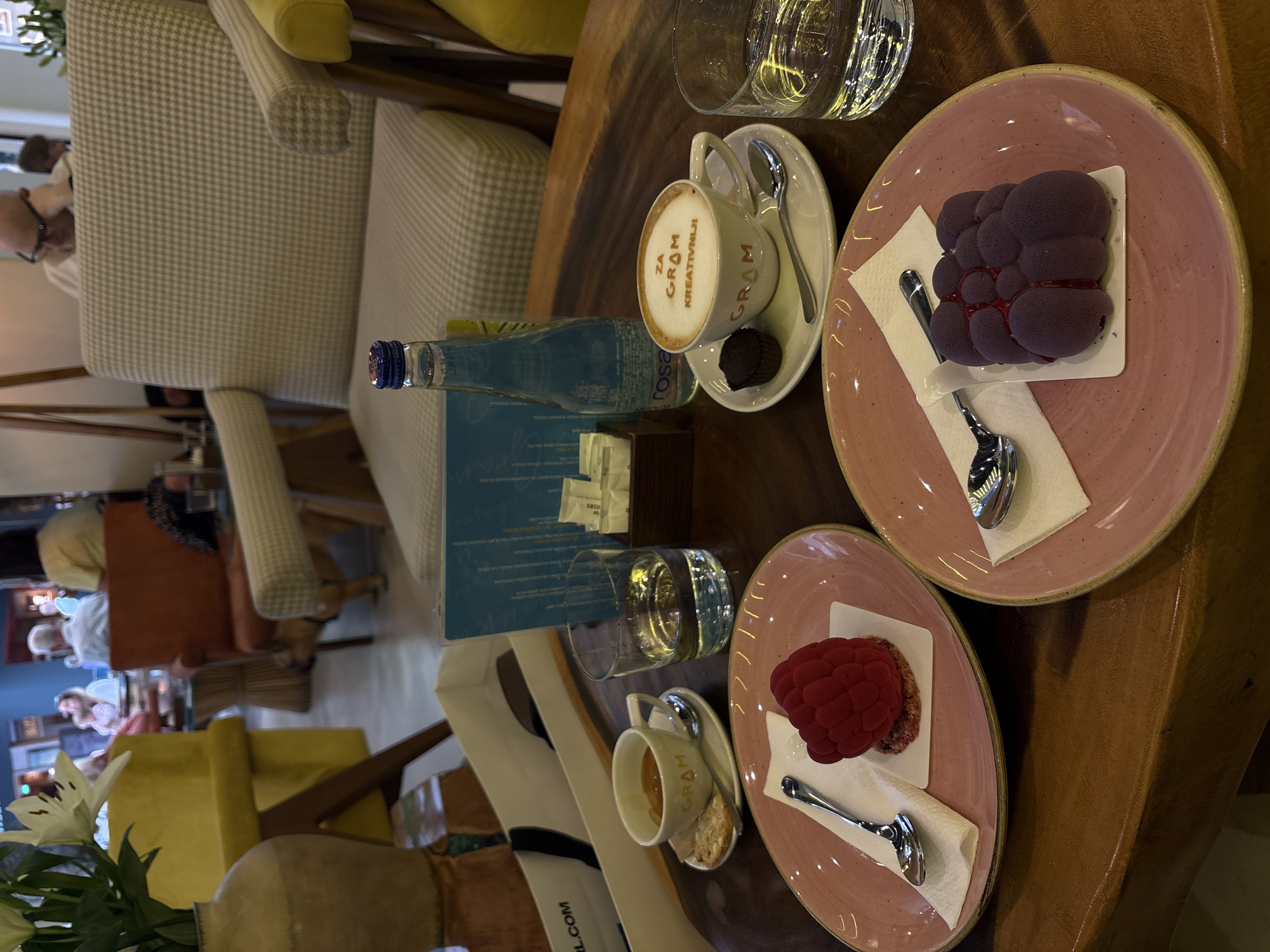 The image depicts a table setting in an indoor establishment, possibly a café or restaurant, with at least two people seated at other tables in the background. The location is suggested to be Belgrade, Serbia, based on the branding present in the photo (e.g., "Za Gram Kreativniji," the "Gram" logo on the coffee cup).
On the table, there are several items arranged:
* Two pink plates, each with a small cake or dessert. One cake is reddish-purple and resembles a cluster of berries, while the other is bright red and round. Each cake sits on a white napkin.
* Two small cups of coffee or espresso, one with a "Gram" logo on the foam, each with a saucer and a small spoon. One cup also has a small chocolate or pastry on the saucer.
* Two glasses of water.
* A blue glass bottle of Rosa water.
* A menu or information card in front of the water bottle.
* Packets of sugar or sweetener.
* White flower arrangements.
The table appears to be made of wood with a natural finish. Around the table are chairs, including one upholstered in a beige and white patterned fabric and another with a yellow cushion. The lighting appears to be natural daylight. Other customers are seated and visible in the background.