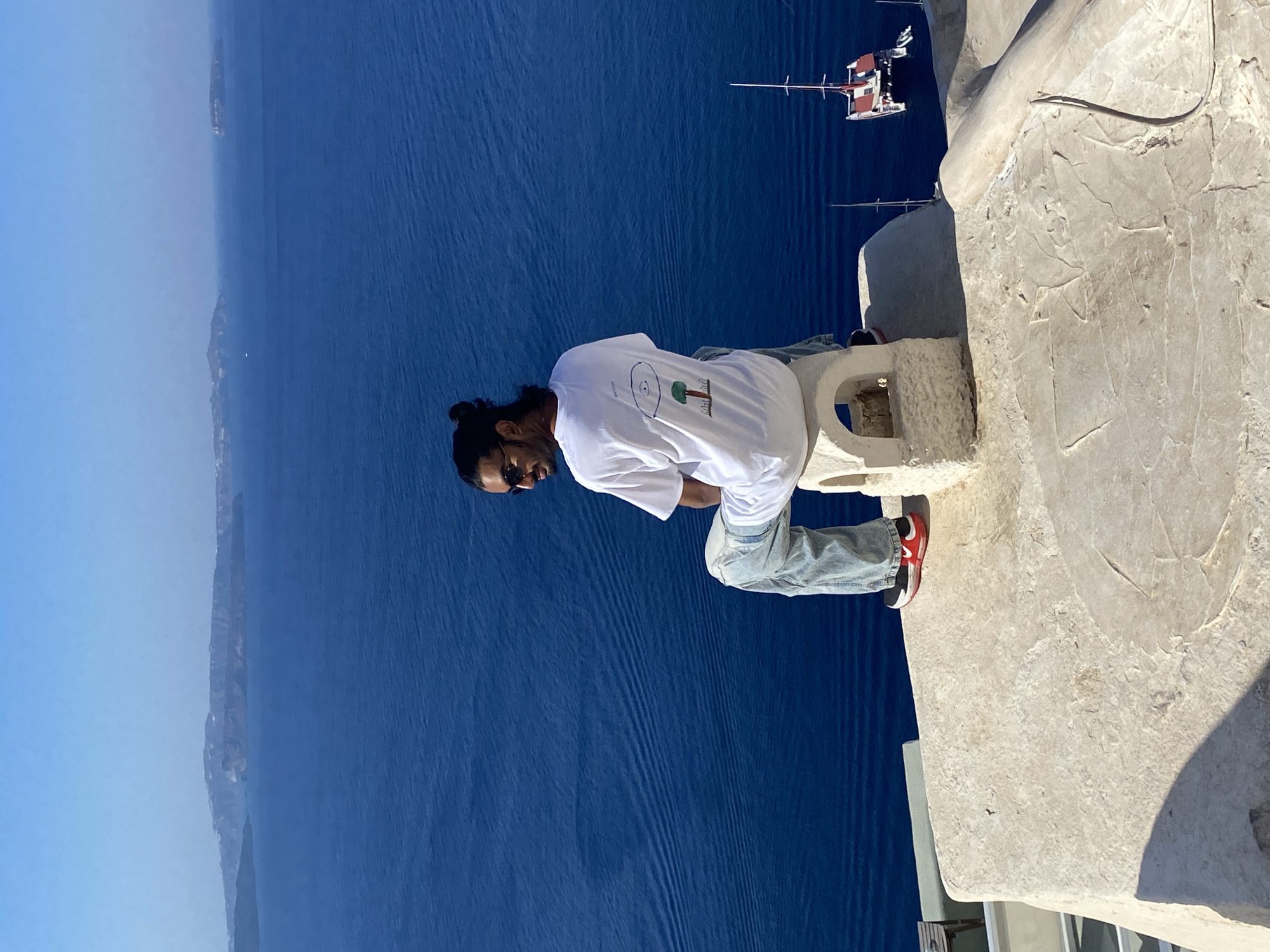 A man with dark hair pulled back in a bun and sunglasses is sitting on a stone structure, facing away from the camera and looking out over the Aegean Sea. He is wearing a white t-shirt with a graphic print on the back and light-wash jeans. His feet are resting on the stone platform. In the background, the deep blue sea stretches to the horizon, meeting a clear, light blue sky. Distant islands or landmasses can be seen along the horizon. A small red and white boat with two masts is docked in the water to the right of the frame. The foreground shows a textured, light-colored stone surface, part of a rooftop or patio. The scene appears to be in Ia, a municipal unit of the island of Santorini, Greece, known for its stunning caldera views.