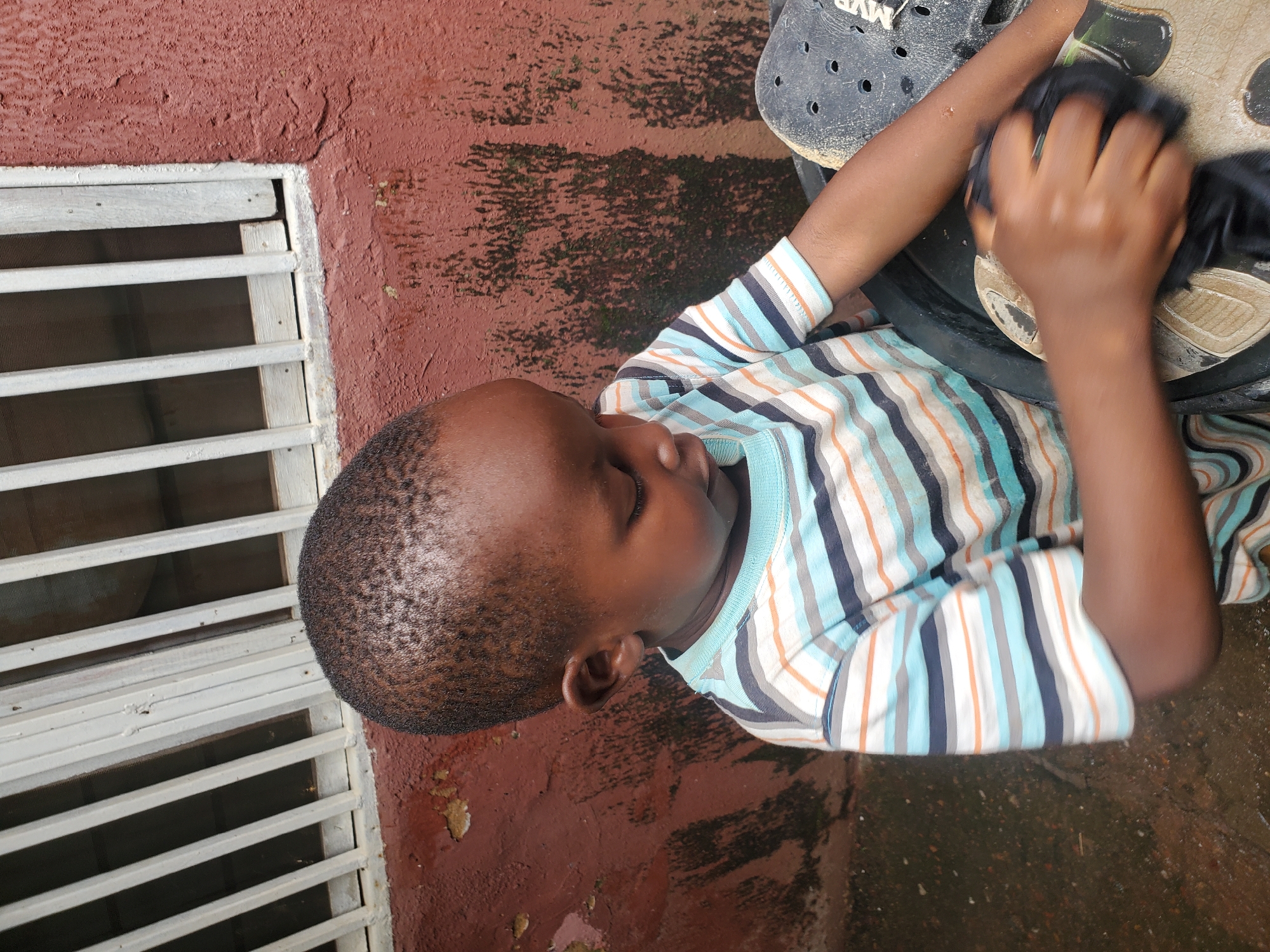 A young child, likely a boy, is shown from a high angle, lying on a reddish-brown textured ground that appears to be outdoors. The child is wearing a striped t-shirt with horizontal bands of light blue, white, dark blue, and orange. His eyes are closed, and his head is tilted downwards, suggesting he is asleep or resting. His hands are occupied with a dark-colored object, possibly a shoe or a piece of fabric, which he is holding and touching with his right hand.
To the left of the child, there is a section of white slatted window bars against a reddish wall. The ground surface has areas of dark discoloration, possibly from water or dirt. Partially visible in the upper right corner is a black object with circular holes, resembling a clog-style shoe. The lighting suggests it is daytime, and there are no clear indications of the weather or specific time of day, other than it being outdoors. The overall scene is casual and appears to be in a residential area. No visible text is present in the image.