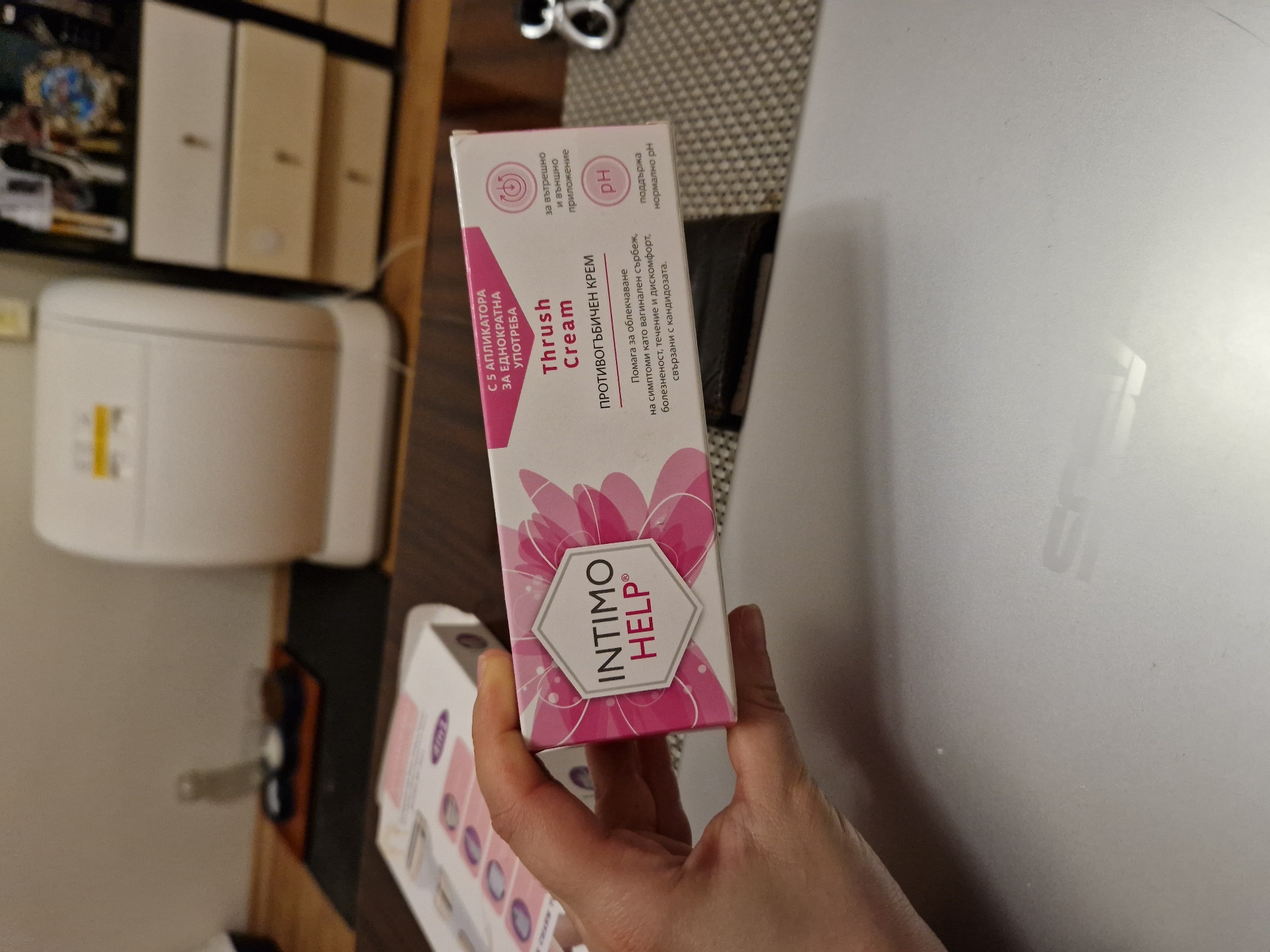 A person is holding a box of "Intimo Help" Thrush Cream. The box is pink and white with the words "Thrush Cream" written on it in English and Bulgarian. The box also has the logo "Intimo Help" in a white hexagon on a pink floral background. The text indicates that the product is used for internal and external application and helps relieve symptoms like vaginal itching, pain, discharge and discomfort associated with candida. The text also states there are 5 single-use applicators and that it maintains normal pH. The background is a mixture of items and textures, including an object on a wooden shelf and a laptop. The exact location in the city of Lovech, Bulgaria cannot be determined from the image.