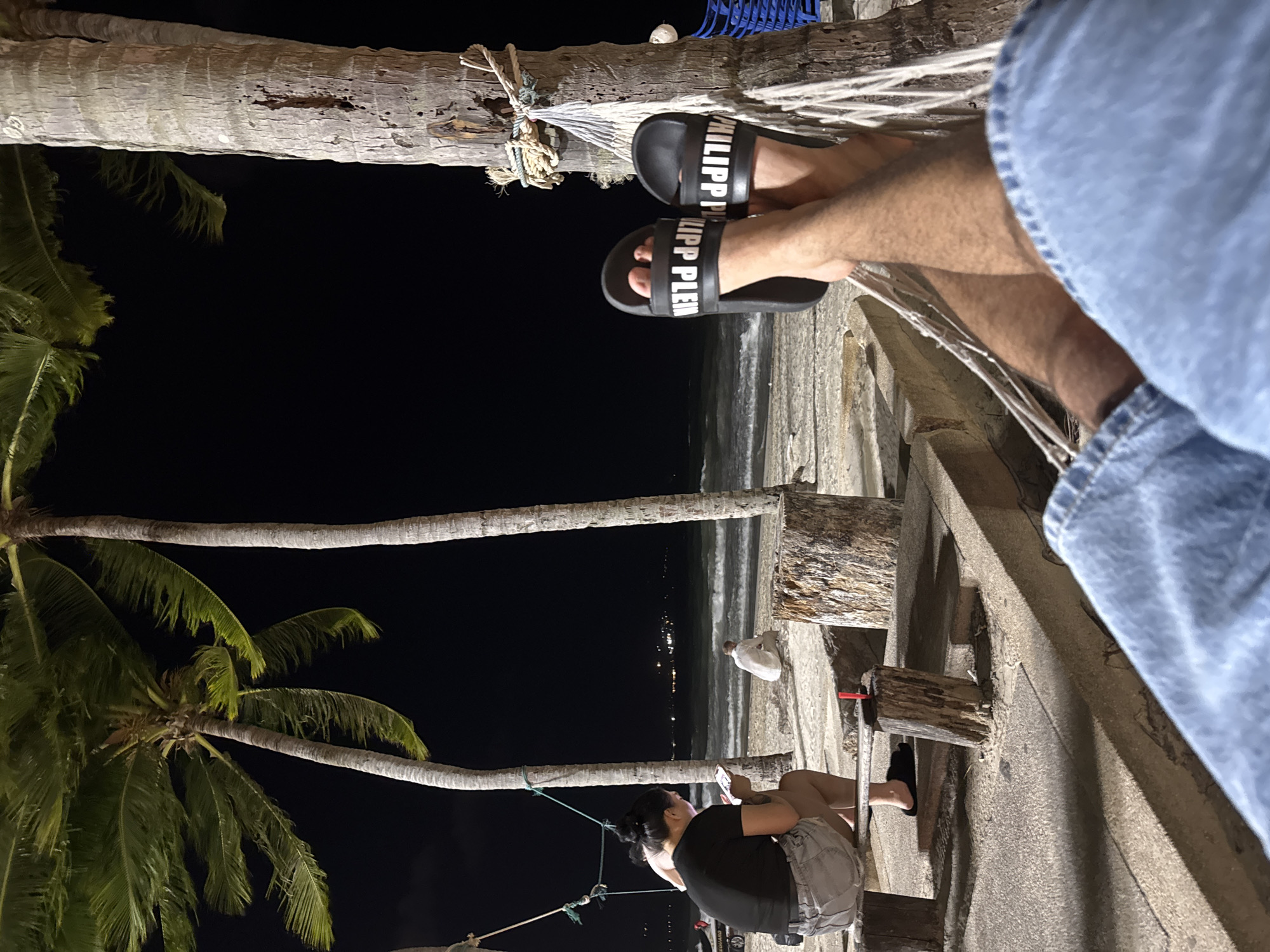 The image captures a nighttime scene in Patong, Thailand. A person wearing Philipp Plein sandals is visible from the knees down, presumably sitting in a hammock strung between palm trees. The hammock is secured with rope. The view from the hammock overlooks a beach at night, with lights visible along the coastline. In the lower portion of the image, another person is seated on what appears to be a wooden structure, possibly using a mobile phone. A white bird stands on the beach in the middle ground. Palm trees are a prominent feature of the scene, framing the dark sky above.