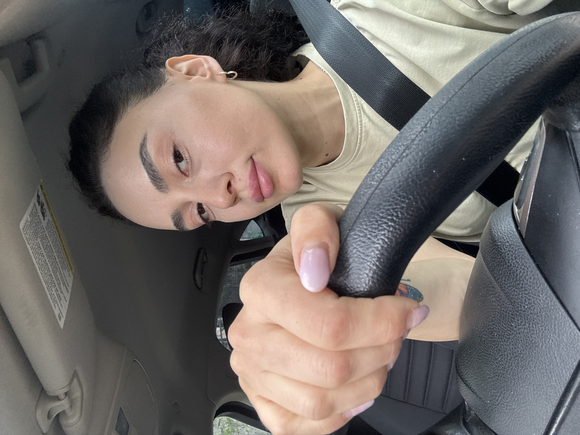 A woman with curly dark hair, fair skin, and light pink nail polish is seated in the driver's seat of a car. She is wearing a beige shirt and a black seatbelt. Her hands are on the black steering wheel. She has an earring in her left ear. A butterfly tattoo is visible on her arm. The car's interior includes a light-colored sun visor. The location is unknown and there is no information in the photo indicating that it is Tbilisi, Georgia.