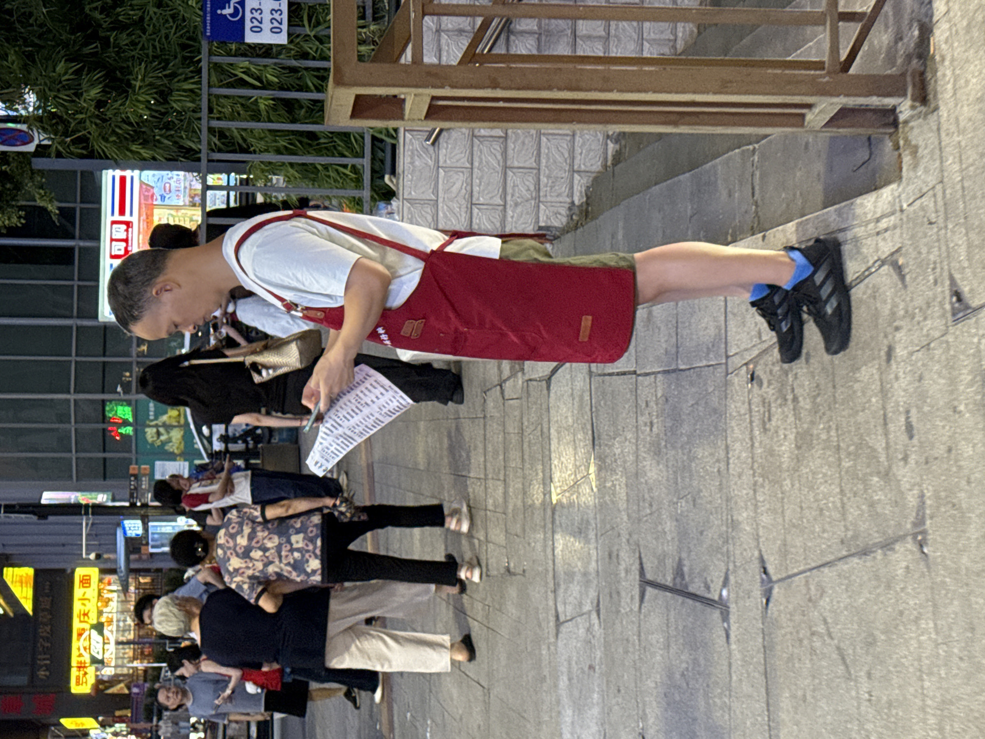 A man wearing a red apron over a white t-shirt and olive green shorts stands on a sidewalk in Yuzhong District, China, holding a piece of paper and a mobile phone. He is looking down at the paper, appearing to be reading it. The man is wearing black athletic shoes with blue socks. Behind him, several other people are walking on the sidewalk. To the right, a metal railing and a bamboo plant are visible. In the background, there are buildings with illuminated signs, typical of a bustling urban environment. The lighting suggests it is evening or night.