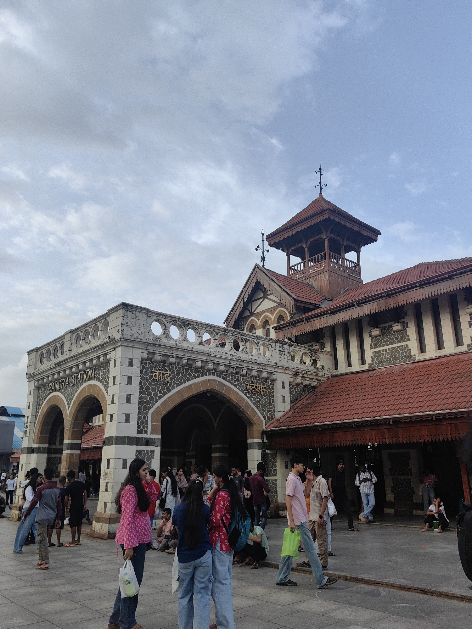 The image shows the exterior of Bandra Station, a railway station in Mumbai, India. The station features distinct architecture with arches, stone cladding, and red tiled roofs, including a prominent wooden tower. A group of people are gathered in front of the station, some walking, some standing, and some sitting. The sky is partly cloudy. The individuals are dressed in casual clothing. The scene appears to be a public space during the daytime.