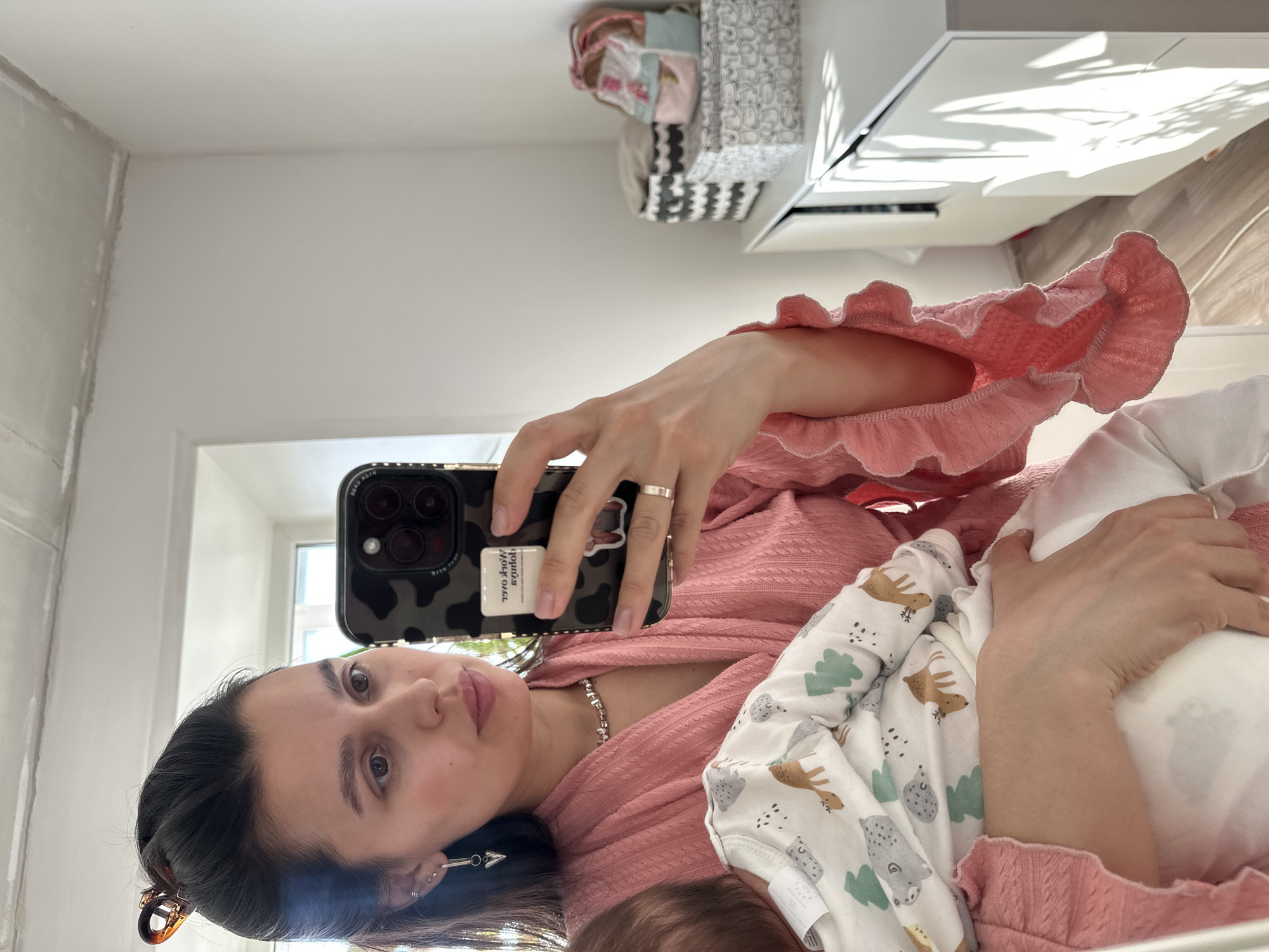 A woman is holding a baby and taking a selfie. She is wearing a pink ruffled top and a silver necklace. She has a silver ring on her left ring finger. Her phone has a black-and-white camouflage case. The baby is wearing a white onesie with images of trees and woodland creatures. The background includes a white wall, a window, a white dresser, and a storage box. There is no explicit evidence in the image indicating that the location is in the city of Chaikovsky, Russia.