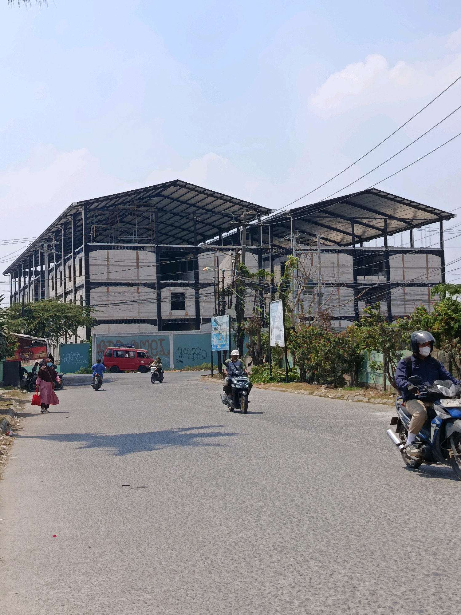 In Babelan, Indonesia, a two-story building under construction dominates the background. The structure features a metal frame with visible concrete blocks and is partially covered by a corrugated metal roof. The building appears to be situated along a paved road where several individuals are operating motorcycles. In the foreground, a person wearing a mask and a dark jacket rides a motorcycle. To the left, another person, also wearing a mask and a pink dress, walks on the side of the road, carrying a red bag. A red van is parked on the street, and further back, more motorcycles are visible, some with riders. The environment is urban, with trees and greenery lining the street and a clear sky above. Graffiti is present on a wall behind the parked van.