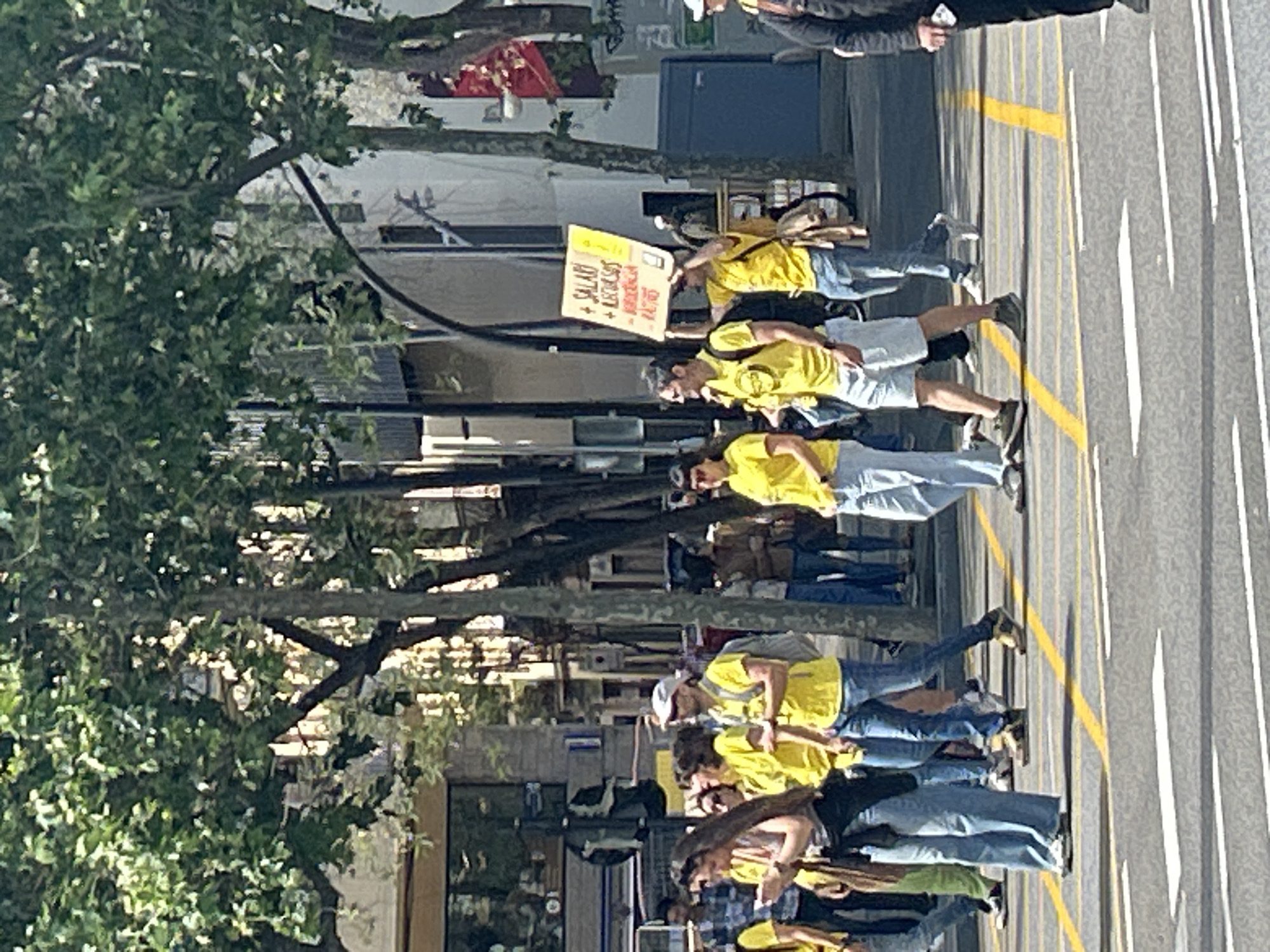A group of people wearing yellow shirts are walking along a street in Barcelona, Spain. They are carrying a sign that reads "SALARI + LLEGISLABES DIGNES". The street is marked with yellow lines. There are trees lining the street. Buildings are visible in the background.